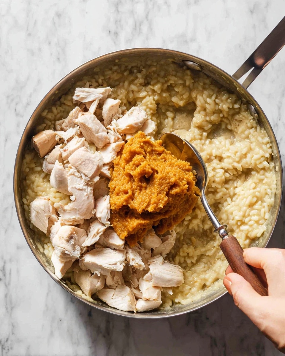 In a metal pan, there is a creamy light beige risotto forming the base layer, with soft, well-cooked rice grains visible. On top of the risotto, chopped pieces of cooked chicken with a white and slightly brown outer texture are scattered evenly. Above the chicken, there is a thick dollop of orange-brown pumpkin or squash puree placed in the center. A silver spoon with a wooden handle is held by a woman's hand, scooping some of the puree over the chicken. The pan rests on a white marbled surface. photo taken with an iphone --ar 4:5 --v 7
