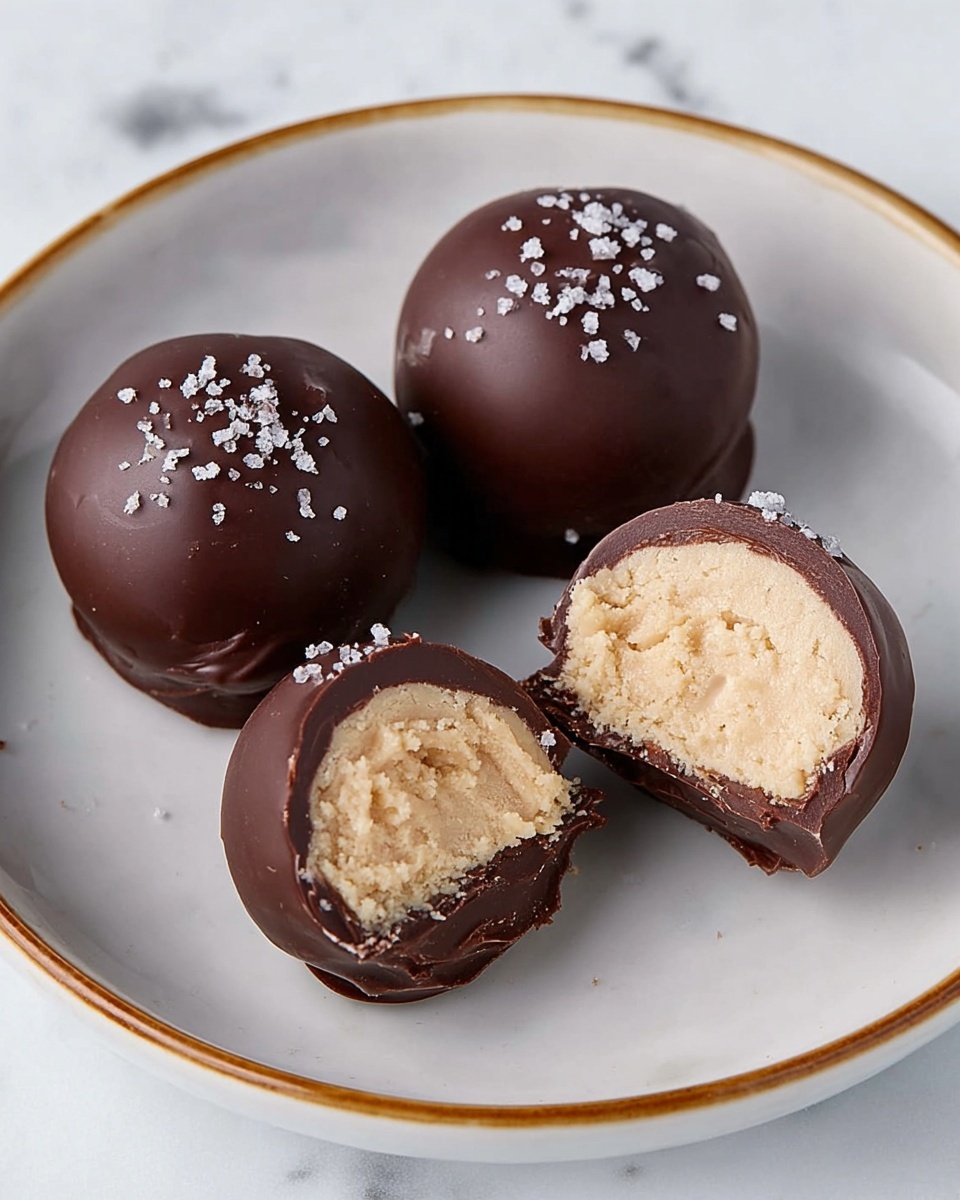 The image shows four round chocolate balls arranged on a white plate with a gold rim, set on a white marbled surface. Three of the balls are fully covered in smooth, dark chocolate with small white salt flakes sprinkled on top, giving a slightly rough texture. The fourth ball is cut in half, revealing a thick, creamy, light beige filling with a soft, crumbly texture inside, surrounded by a thin layer of dark chocolate shell. The chocolate shell is shiny and smooth, contrasting with the matte, textured filling inside. Photo taken with an iphone --ar 4:5 --v 7