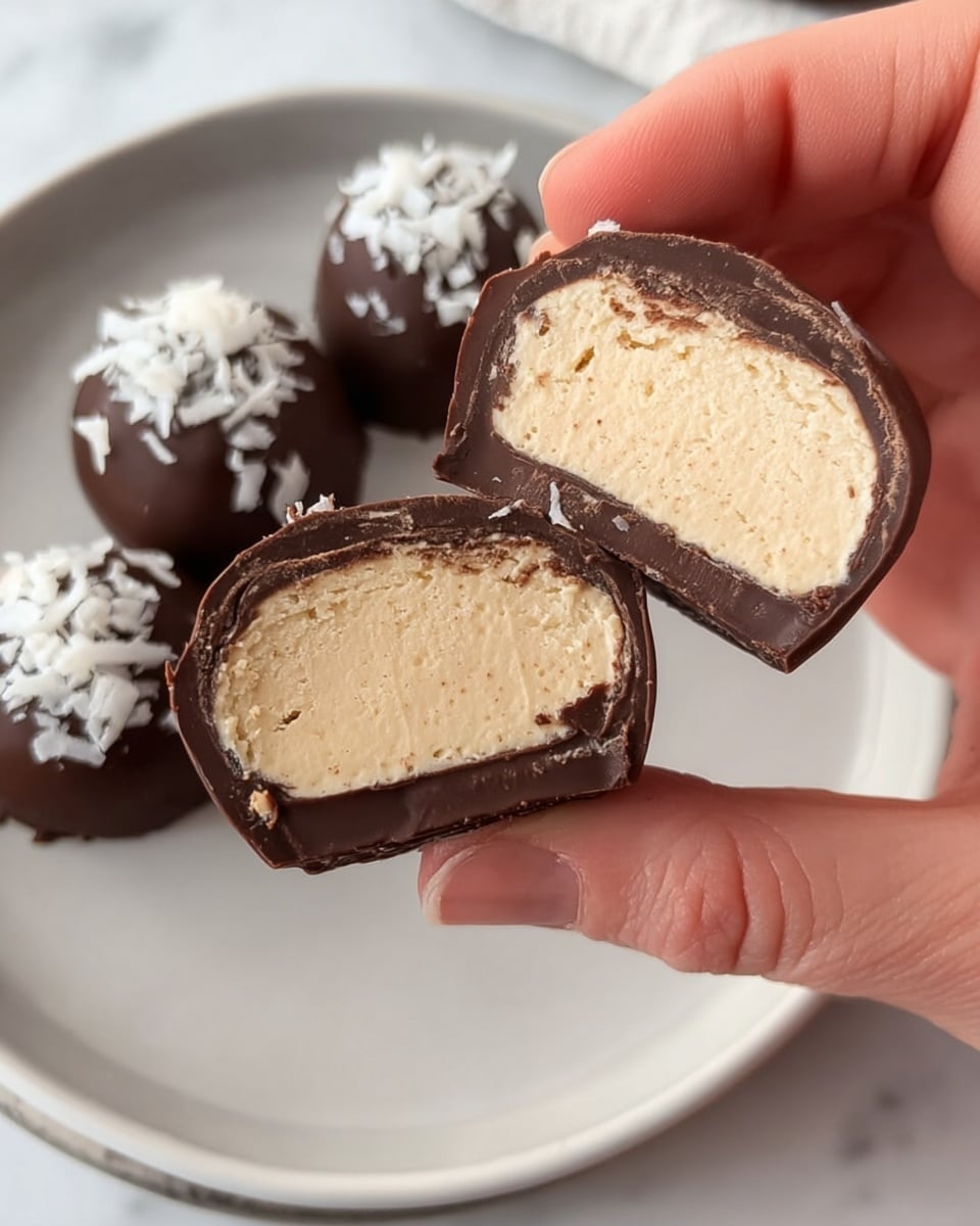 A close-up of a woman's hand holding two pieces of a round chocolate candy cut in half to show the inside. The candy has two layers: a smooth, light beige creamy center with a soft texture, surrounded by a dark brown chocolate shell with a slightly rough edge. The background shows a white plate with three whole round chocolates topped with white flakes, all placed on a white marbled surface. photo taken with an iphone --ar 4:5 --v 7