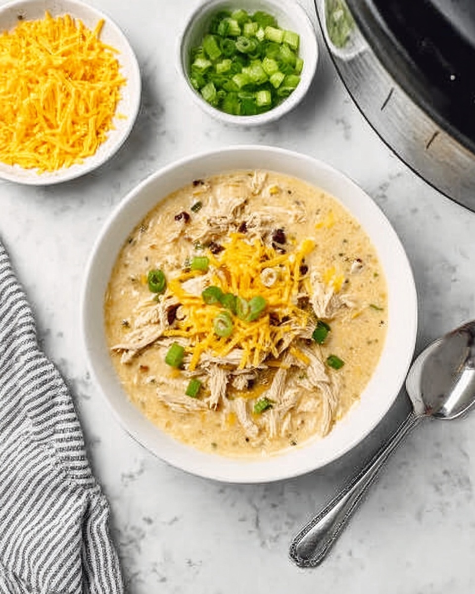 A white bowl filled with a creamy soup that has shredded light brown chicken pieces and melted yellow cheese on top, with small green onion pieces scattered as garnish. The bowl is placed on a white marbled surface near a small white bowl of chopped green onions and another white bowl of grated yellow cheese. A silver spoon lies next to the bowl, and a gray and white striped cloth is partially visible on the left side. photo taken with an iphone --ar 4:5 --v 7