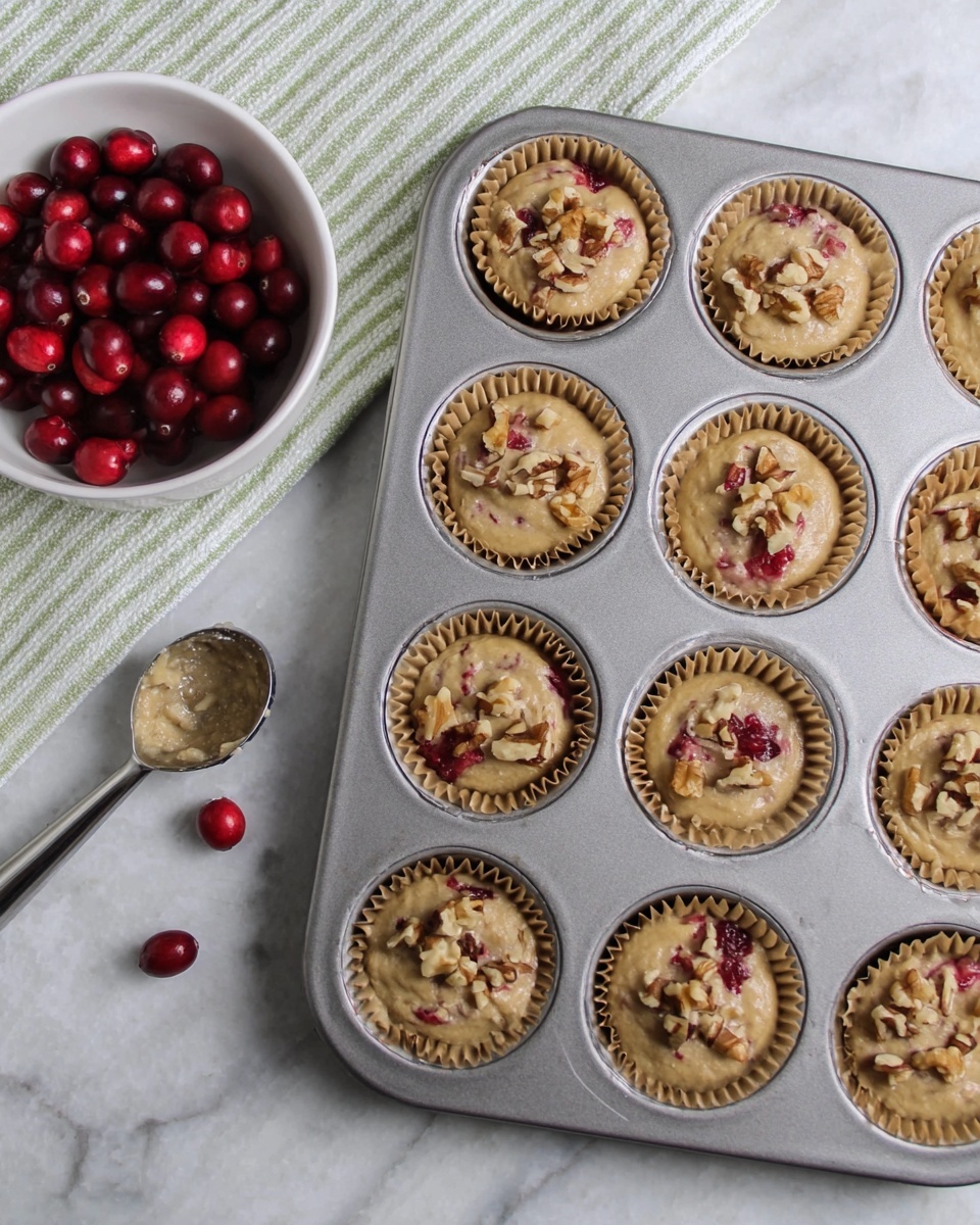 A metal muffin tray with 12 cups lined with brown paper holds a light beige batter mixed with red cranberries and topped with small chunks of chopped walnuts. The batter has a smooth texture with visible bits of walnuts and cranberries inside. To the upper left, a white bowl filled with red cranberries sits on a light green and white striped cloth on a white marbled surface. A metal ice cream scoop with some batter on it rests on the surface near three loose cranberries. The scene is well-lit, showing a fresh baking setup photo taken with an iphone --ar 4:5 --v 7