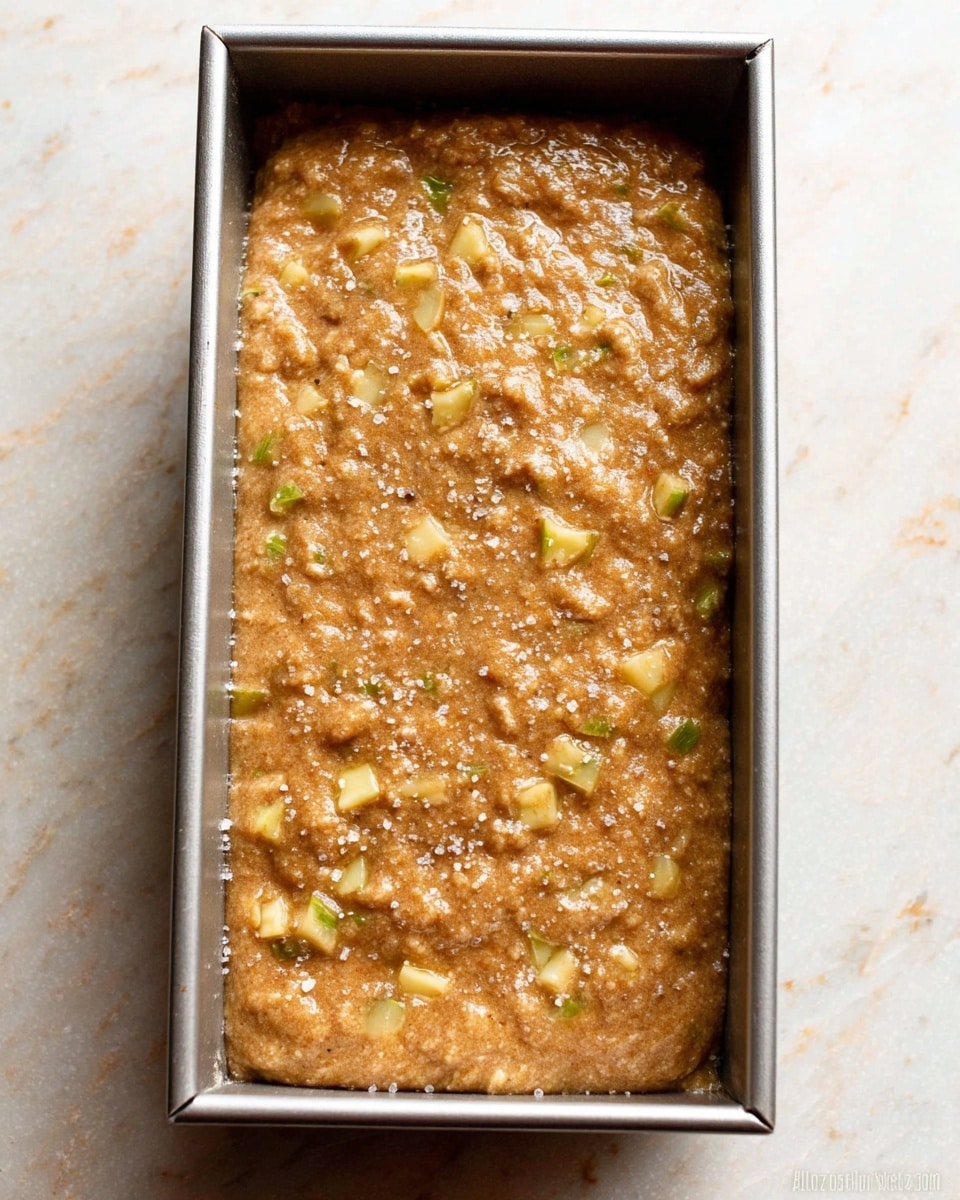 A rectangular silver metal pan filled with a thick, brown batter that has visible chunks of light yellow and green pieces scattered throughout, giving it a textured look. The surface of the batter is sprinkled with a layer of coarse sugar crystals, adding a slight sparkle and roughness to the top. The pan is placed on a white marbled textured surface. photo taken with an iphone --ar 4:5 --v 7