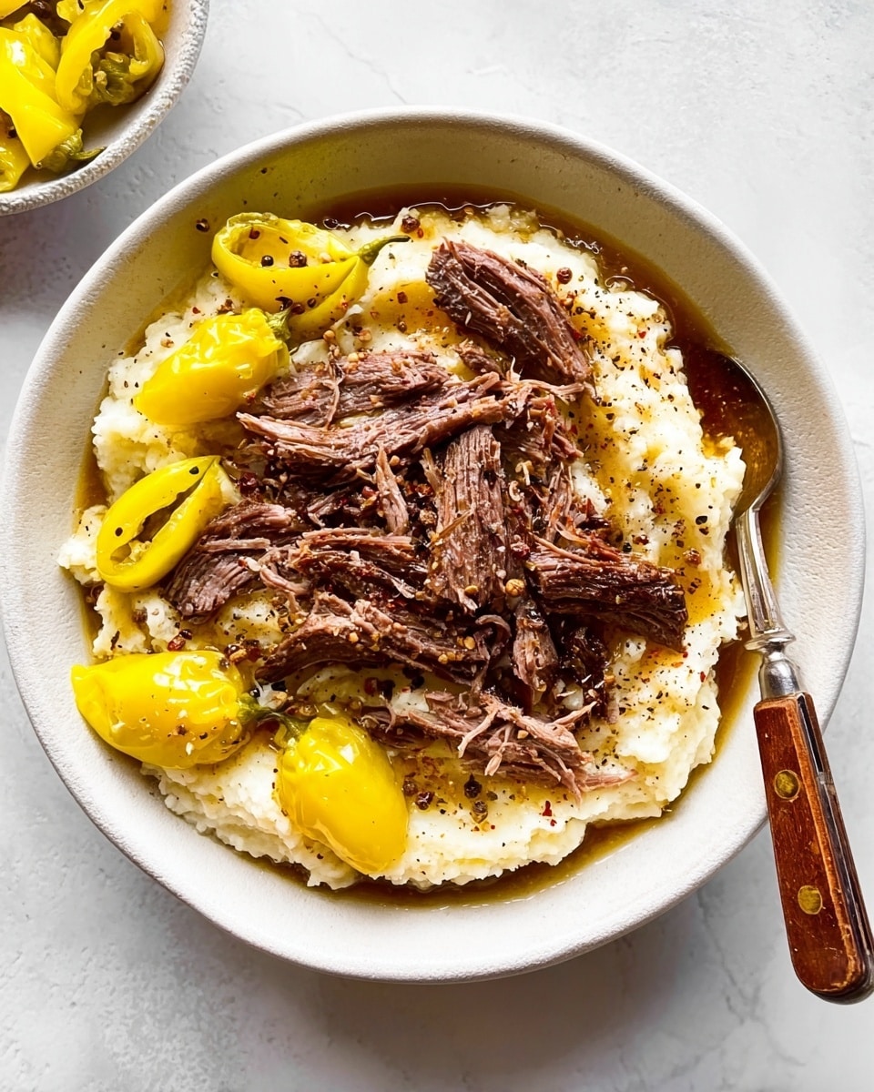 The dish is served in a deep white bowl on a white marbled surface. The bottom layer consists of creamy mashed potatoes with a smooth texture, spread evenly across the bowl. On top of the mashed potatoes, there are several whole yellow pepperoncini peppers placed around the edges, adding a bright yellow color contrast. In the center and on top, there are moist, shredded pieces of dark brown cooked meat, arranged loosely with some meat chunks showing fibers. A fork with a wooden handle is placed inside the bowl on the right side. The overall colors include creamy off-white, bright yellow, and rich dark brown, with some black pepper flakes sprinkled lightly on top. photo taken with an iphone --ar 4:5 --v 7