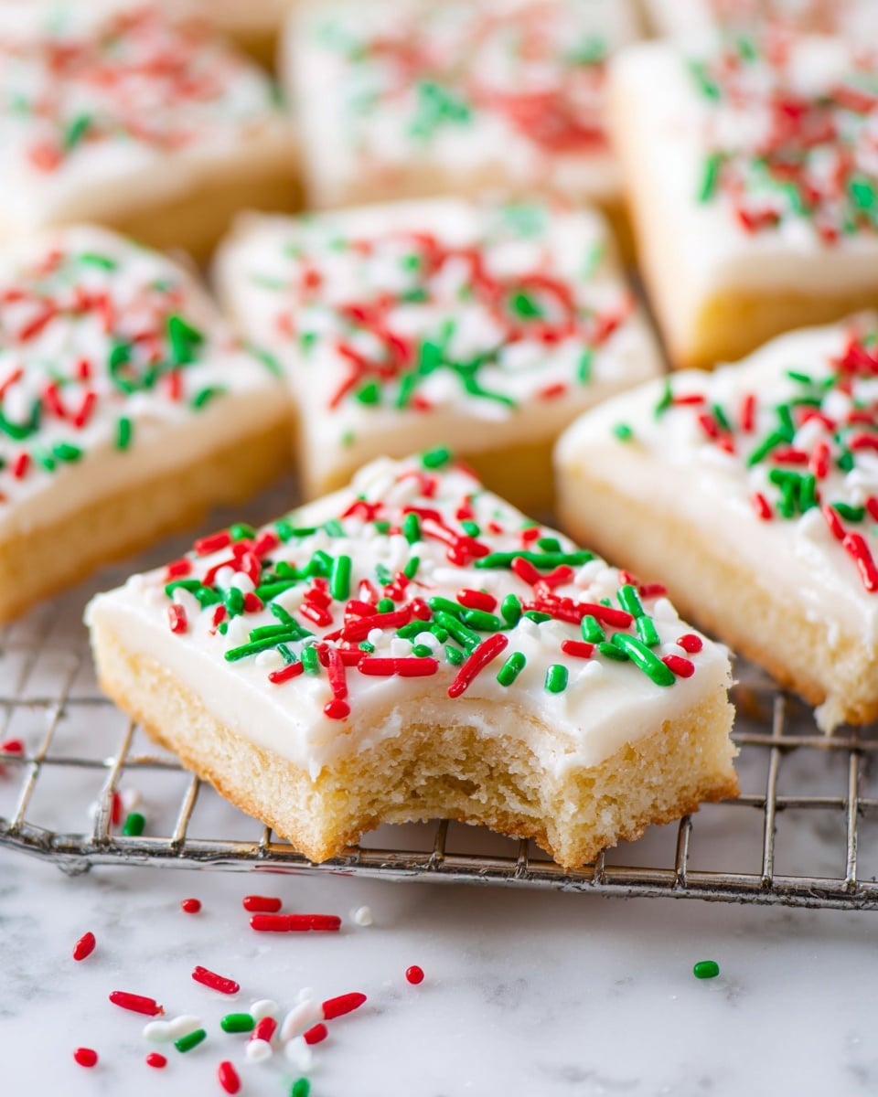 The image shows several square cookies arranged on a metal rack, placed on a white marbled surface. Each cookie has two layers: a light golden tan base with a soft, crumbly texture, and a thick, smooth white frosting on top. The frosting is decorated with red and green elongated sprinkles scattered evenly across the surface. One cookie in the front has a bite taken out of the edge, showing the layers clearly. There are loose sprinkles scattered on the white marbled surface around the cookies. Photo taken with an iphone --ar 4:5 --v 7