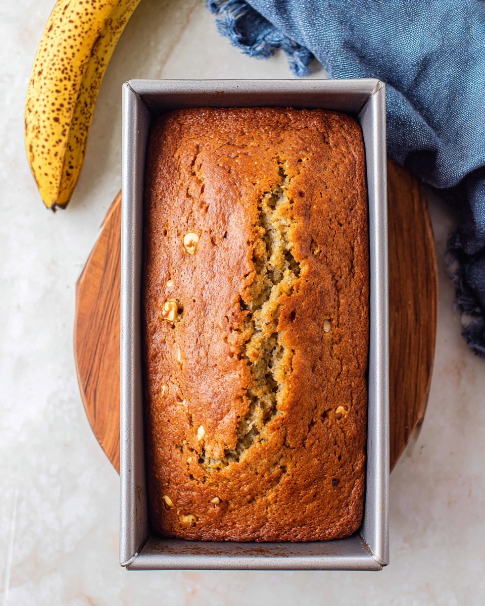 A golden brown loaf of banana bread in a silver rectangular baking pan, with a cracked top center showing a moist, soft inside and small nut pieces scattered throughout the bread’s surface; the pan rests on a wooden round board on a white marbled surface, with a ripe, spotted banana partially visible in the top left corner and a crumpled blue cloth in the top right corner, photo taken with an iphone --ar 4:5 --v 7
