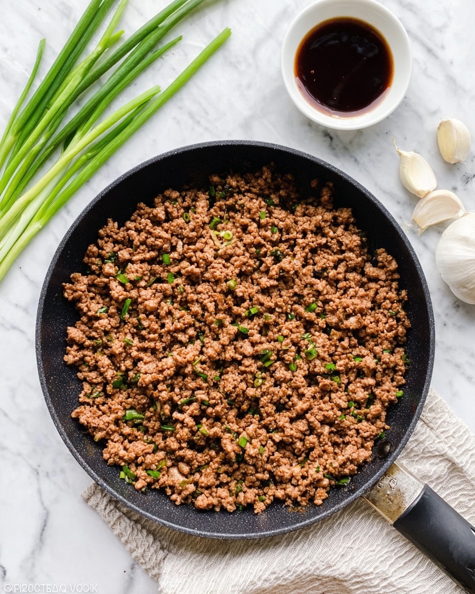 A close-up view of a black frying pan filled with cooked ground meat that is brown with small green herb pieces mixed in, sitting on a white marbled surface with a cloth napkin folded next to it. Above the pan, some green stalks and a small white bowl filled with dark brown sauce are placed, along with two garlic cloves. The pan handle extends to the right, and the scene is bright and clear. photo taken with an iphone --ar 4:5 --v 7