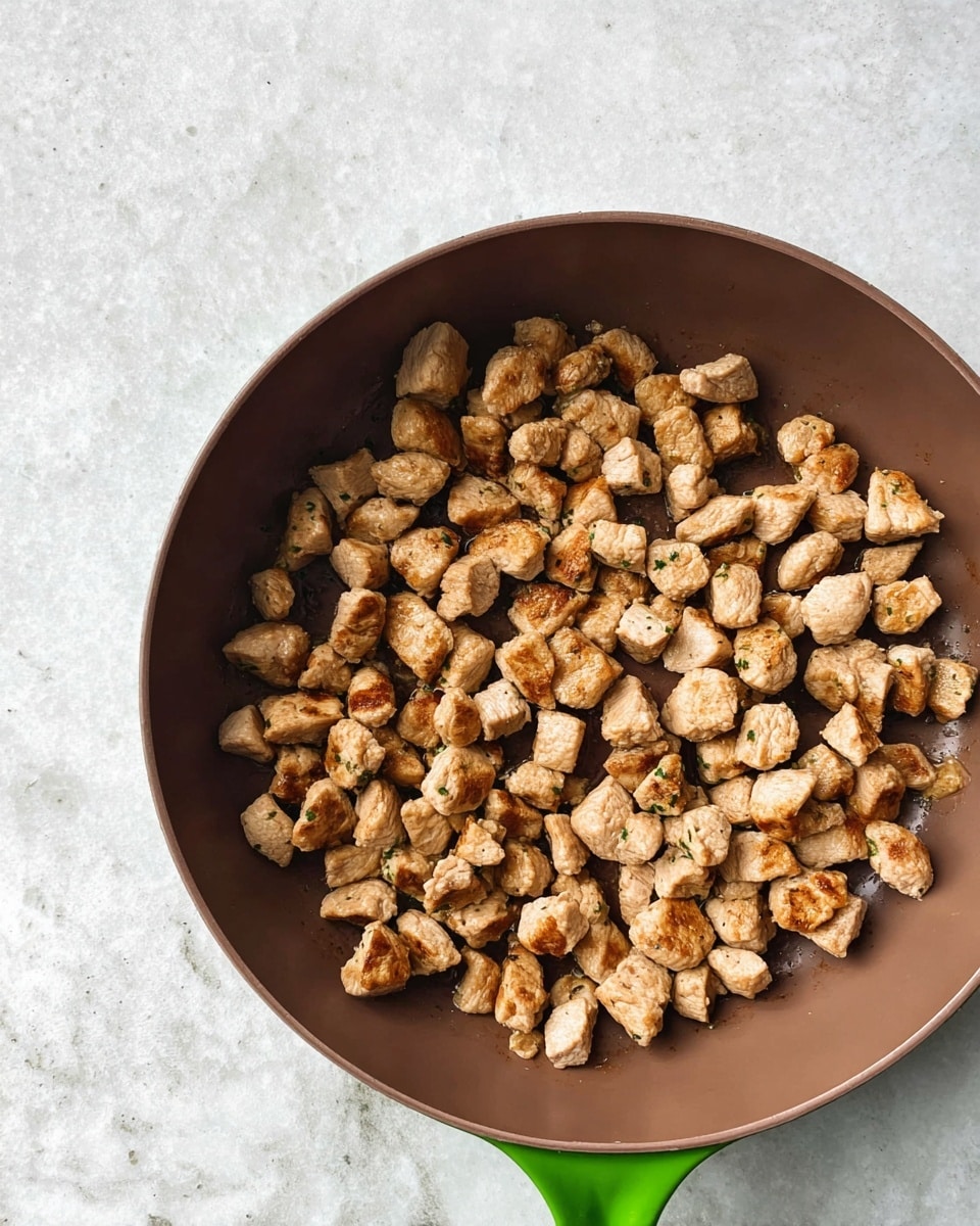 A close-up top view of a green handled brown pan filled with small, cooked, light brown meat pieces scattered evenly across the pan's surface, set against a white marbled background photo taken with an iphone --ar 4:5 --v 7