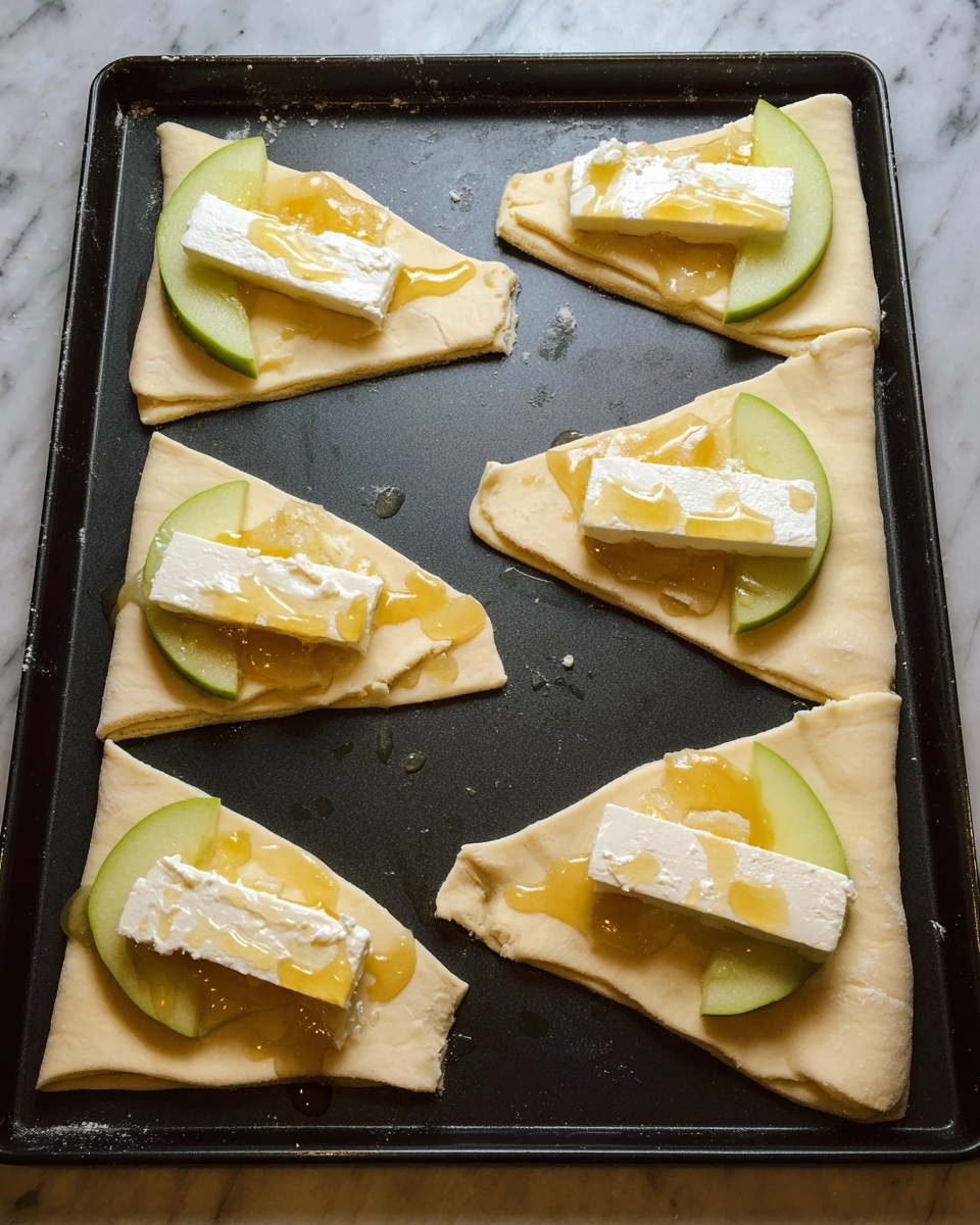 On a dark baking tray, there are seven uncooked triangular pieces of light beige dough spaced apart. At the wide end of each triangle, there is a green apple slice placed flat, topped with a thick, rectangular piece of white soft cheese with a rind. A golden honey-like syrup is drizzled over the cheese and apple, creating small shiny pools around the base of the layers. The dough pieces have a slightly rough texture with uneven edges. The photo is taken against a white marbled surface. Photo taken with an iphone --ar 4:5 --v 7