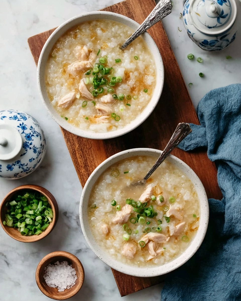 The image shows two white bowls filled with a creamy rice porridge topped with small pieces of tender chicken and sprinkled with finely chopped green onions. Each bowl has a spoon resting inside, partially submerged in the thick, light beige porridge with visible grains of rice and soft chicken chunks. The bowls are placed on a white marbled surface, layered with a wooden board underneath one of the bowls. To the left, there is a small white bowl with more chopped green onions and a small white container with a blue floral pattern. A small wooden bowl with coarse salt and a spoon is also on the surface. A woman's hand is gently holding a blue cloth napkin near the bottom right corner. Photo taken with an iphone --ar 4:5 --v 7