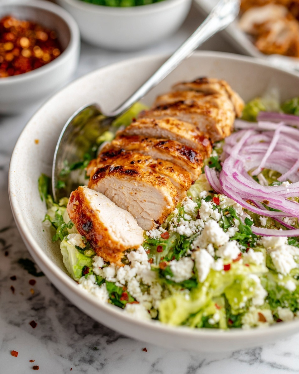 A white bowl sits on a white marbled surface, filled with a layered salad. The bottom layer is finely chopped bright green lettuce, sprinkled lightly with red chili flakes. On top of this, thin slices of raw purple onion rest next to a portion of grilled chicken breast, sliced and arranged in a neat row across the bowl. The chicken has a golden brown, slightly charred outside and a juicy white inside. Crumbled white cheese and chopped green herbs are scattered over the chicken and onions, adding some texture and color. A metal spoon is placed inside the bowl, partially under the chicken. In the background, blurred white bowls with red and green toppings are visible. Photo taken with an iphone --ar 4:5 --v 7