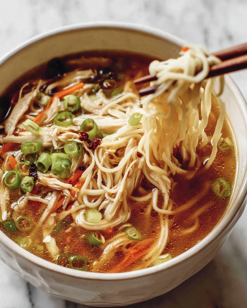 A white bowl filled with clear brown broth with thin white noodles lifted by chopsticks near the top right; in the broth there are shredded light brown chicken pieces, slices of green onion, thin orange carrot strips, and small dark red chili flakes scattered around. The bowl is placed on a white marbled surface. photo taken with an iphone --ar 4:5 --v 7