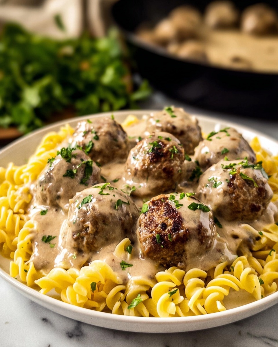 A white plate holds a bed of yellow curly pasta as the first layer, with six brown meatballs covered in creamy light beige sauce placed on top in the second layer. The meatballs have some green herb bits sprinkled on them. The sauce looks smooth and shiny, spreading over both the meatballs and pasta slightly. In the background, out of focus, there is a dark pan and some green leafy herbs on a white marbled surface. Photo taken with an iphone --ar 4:5 --v 7