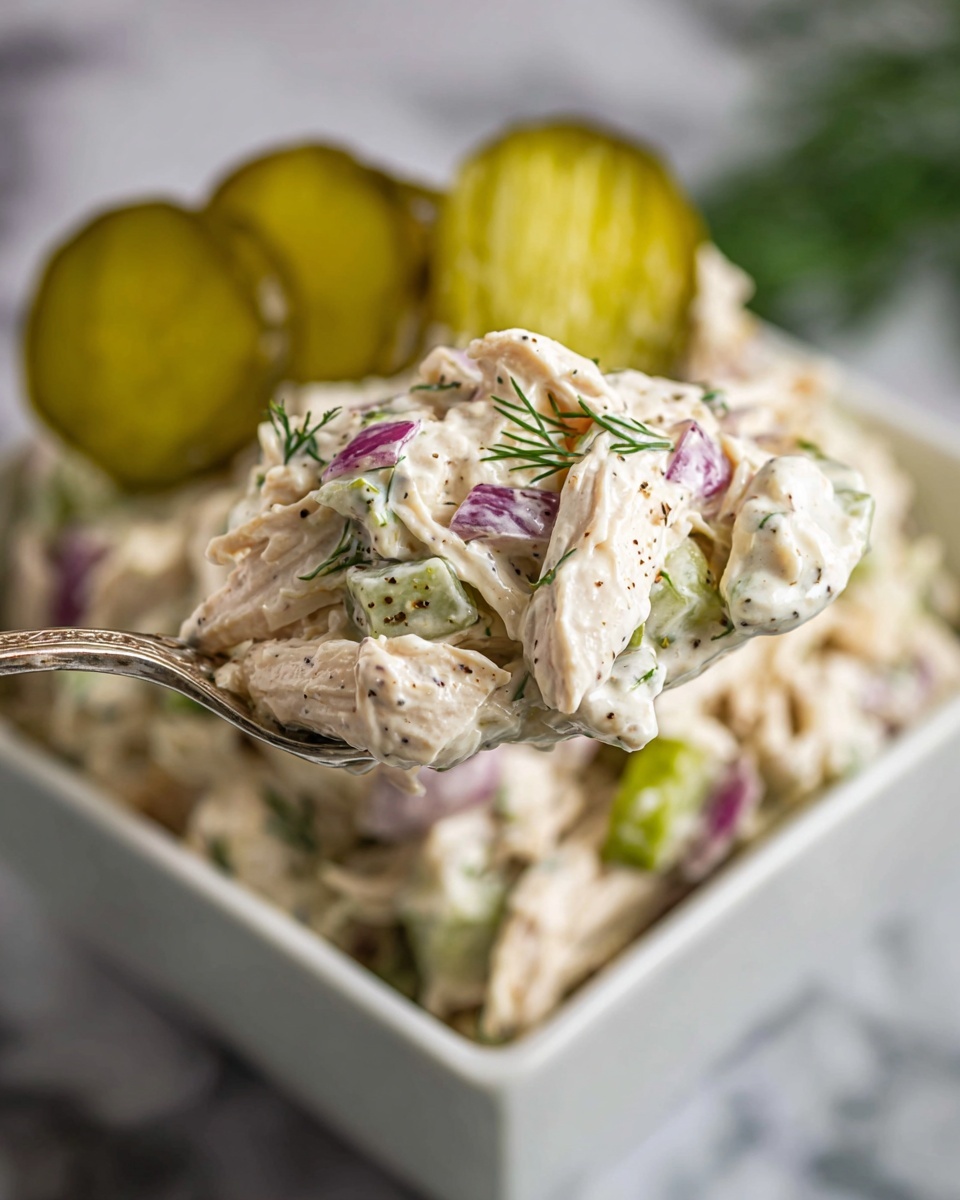 The image shows a close-up of a spoonful of creamy chicken salad held above a white square bowl. The salad consists of shredded white chicken mixed with small pieces of purple-red onion, bits of green celery, and fresh green dill herbs, all coated in a thick, creamy dressing speckled with black pepper. In the background, the bowl is filled with more of the chicken salad and topped with several round light green pickle slices. The setting is on a white marbled surface. photo taken with an iphone --ar 4:5 --v 7