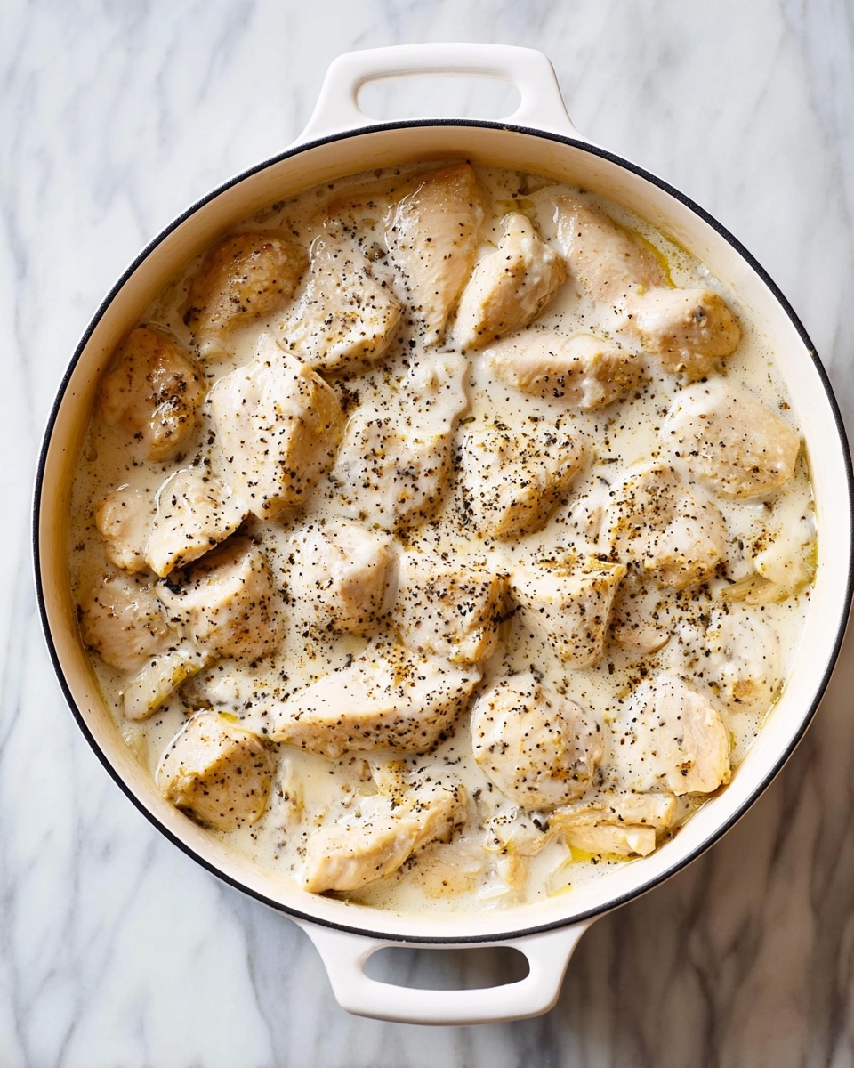 A white oval pot with two handles contains many pieces of light beige chicken covered in a creamy, white sauce with visible black pepper specks scattered on top. The pieces have a soft texture, partly submerged in the thick sauce. The pot sits on a white marbled surface, which adds a clean and bright background to the image. photo taken with an iphone --ar 4:5 --v 7