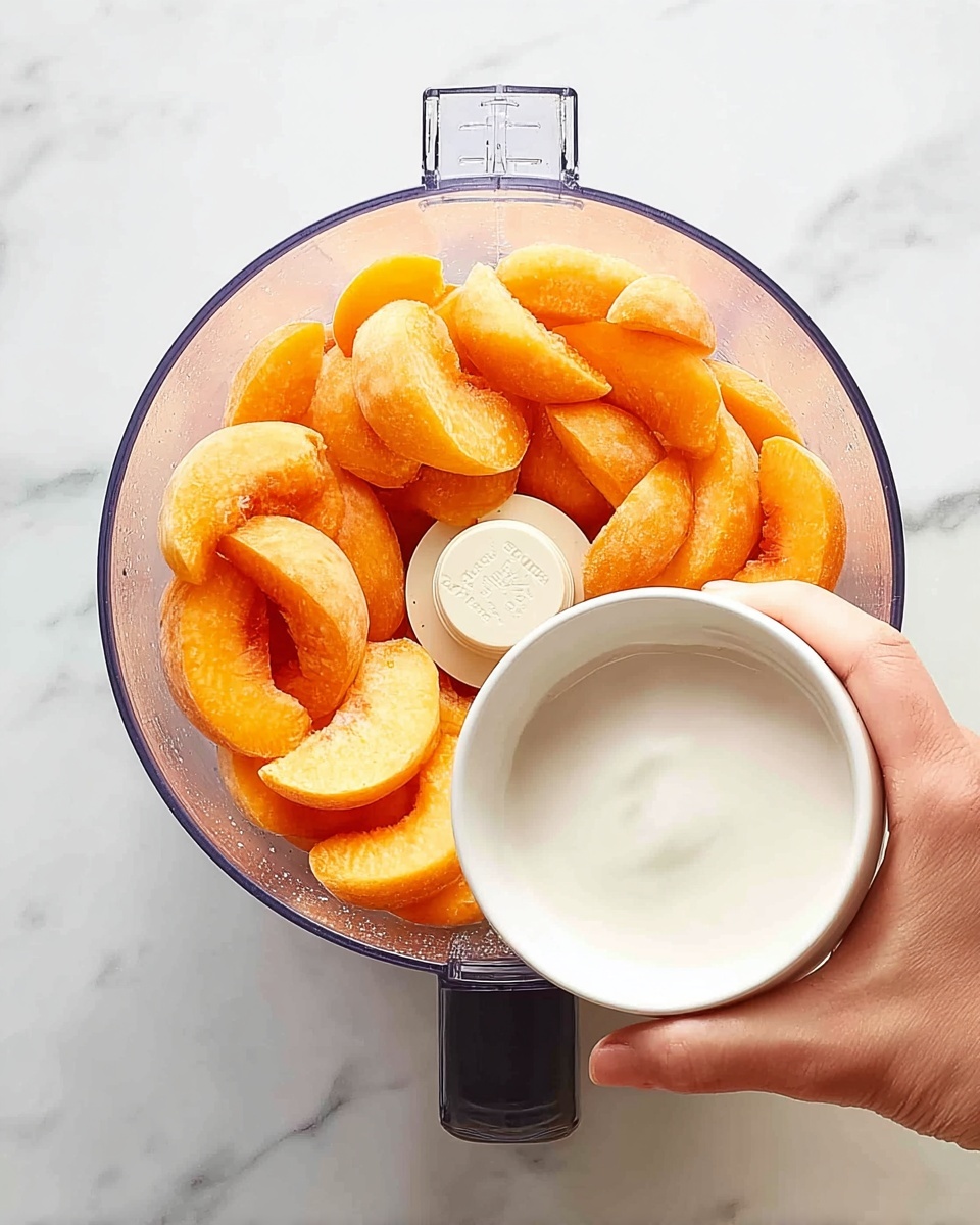 A clear food processor bowl filled with bright orange peach slices, arranged evenly inside the bowl, is shown from above. A woman's hand is holding a small white bowl filled with a smooth white liquid, positioned over the food processor as if about to pour the liquid in. The scene is set on a white marbled surface that adds a clean, fresh background to the image. photo taken with an iphone --ar 4:5 --v 7