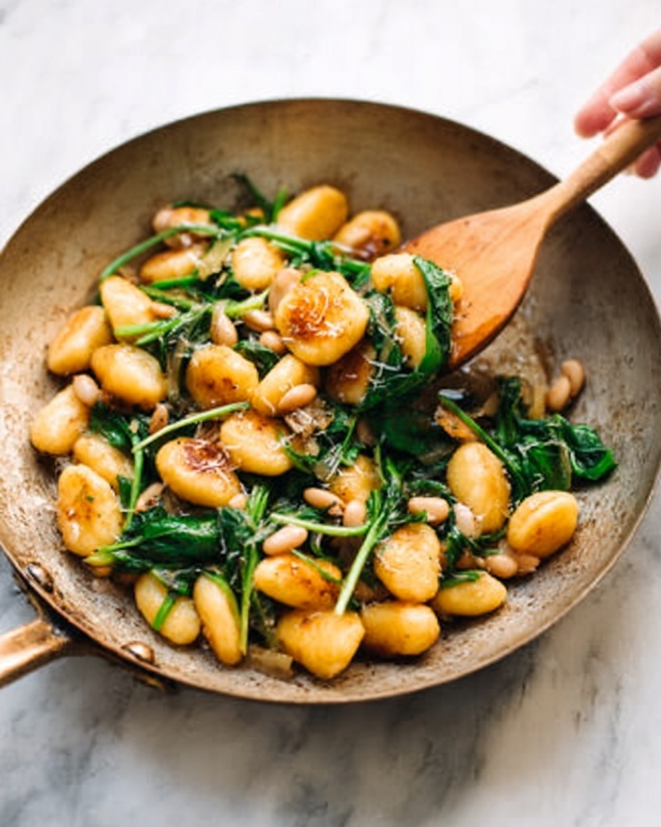 The image shows a metal pan filled with golden-brown gnocchi mixed with bright green spinach leaves and small white beans. The dish looks cooked in olive oil with some light seasoning visible on the gnocchi. A wooden spoon held by a woman's hand is stirring the food, lifting some pieces above the rest. The pan sits on a white marbled surface with soft natural lighting highlighting the textures. Photo taken with an iphone --ar 4:5 --v 7
