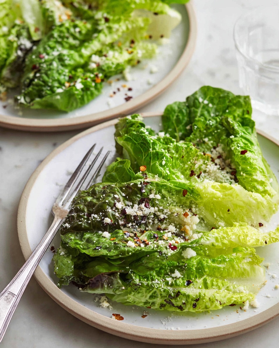 The image shows two white plates on a white marbled surface, each with a small pile of fresh lettuce leaves in different shades of green, including light green and dark green. The leaves have a slightly ruffled texture and are sprinkled with finely grated white cheese and small red chili flakes. On the front plate, a silver fork rests at the bottom left edge, its handle pointing outward. The background plate is slightly out of focus, emphasizing the front plate with the salad. photo taken with an iphone --ar 4:5 --v 7