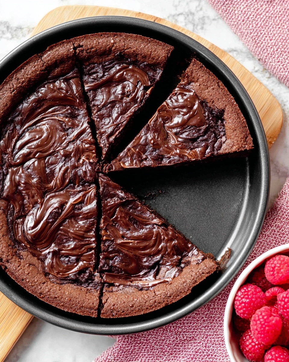 The image shows a round chocolate dessert with two slices cut out, sitting inside a dark baking pan on a white marbled surface. The dessert has one thick layer with a rich, glossy dark chocolate swirl pattern on top of a matte, tender chocolate base that looks soft and slightly cracked. The outer crust is firm and darker brown, forming a raised edge around the dessert. In the lower right corner, there is a white bowl filled with bright red raspberries, adding a pop of color. The scene includes a pink textured cloth beneath the baking pan and part of a woman's hand barely visible holding the pan edge. Photo taken with an iphone --ar 4:5 --v 7