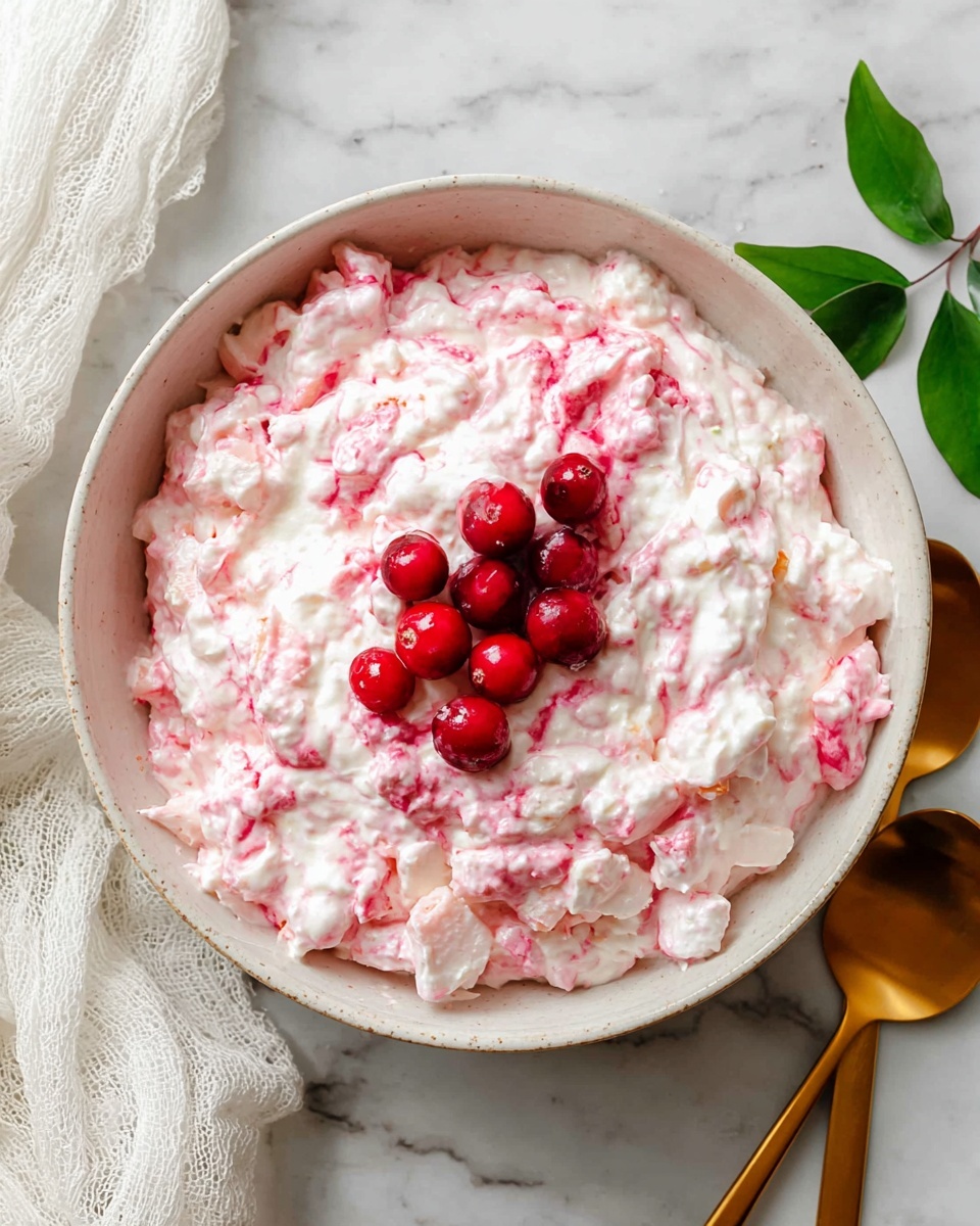 The image shows a white bowl full of a creamy pink salad with visible small white marshmallows and chopped pecans mixed in. Three whole red cranberries sit on top as decoration. The bowl is placed on a white marbled surface with a red and white checkered cloth behind it. More cranberries are scattered in the background around the bowl. A woman's hand is about to pick some from the bowl. photo taken with an iphone --ar 4:5 --v 7