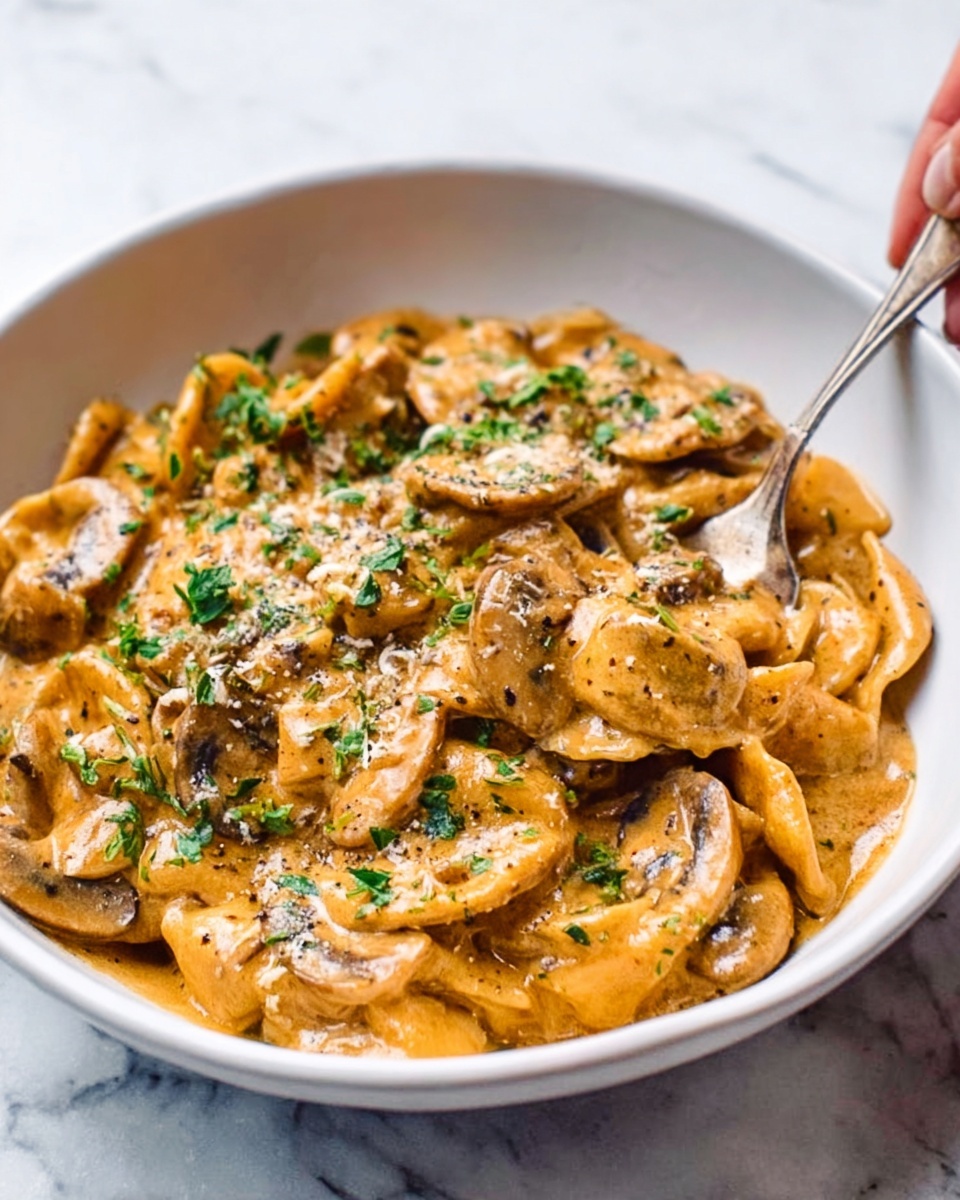 A close-up view of a white bowl filled with several layers of creamy mushroom stroganoff. The bottom layer shows tender mushroom slices in a smooth, orange-brown sauce that looks rich and thick. Small green parsley leaves are lightly sprinkled on top, adding a fresh touch of color. A spoon is partially inside the bowl, with a woman's hand barely touching it on the side. The bowl is placed on a white marbled surface, giving a clean and simple background. photo taken with an iphone --ar 4:5 --v 7