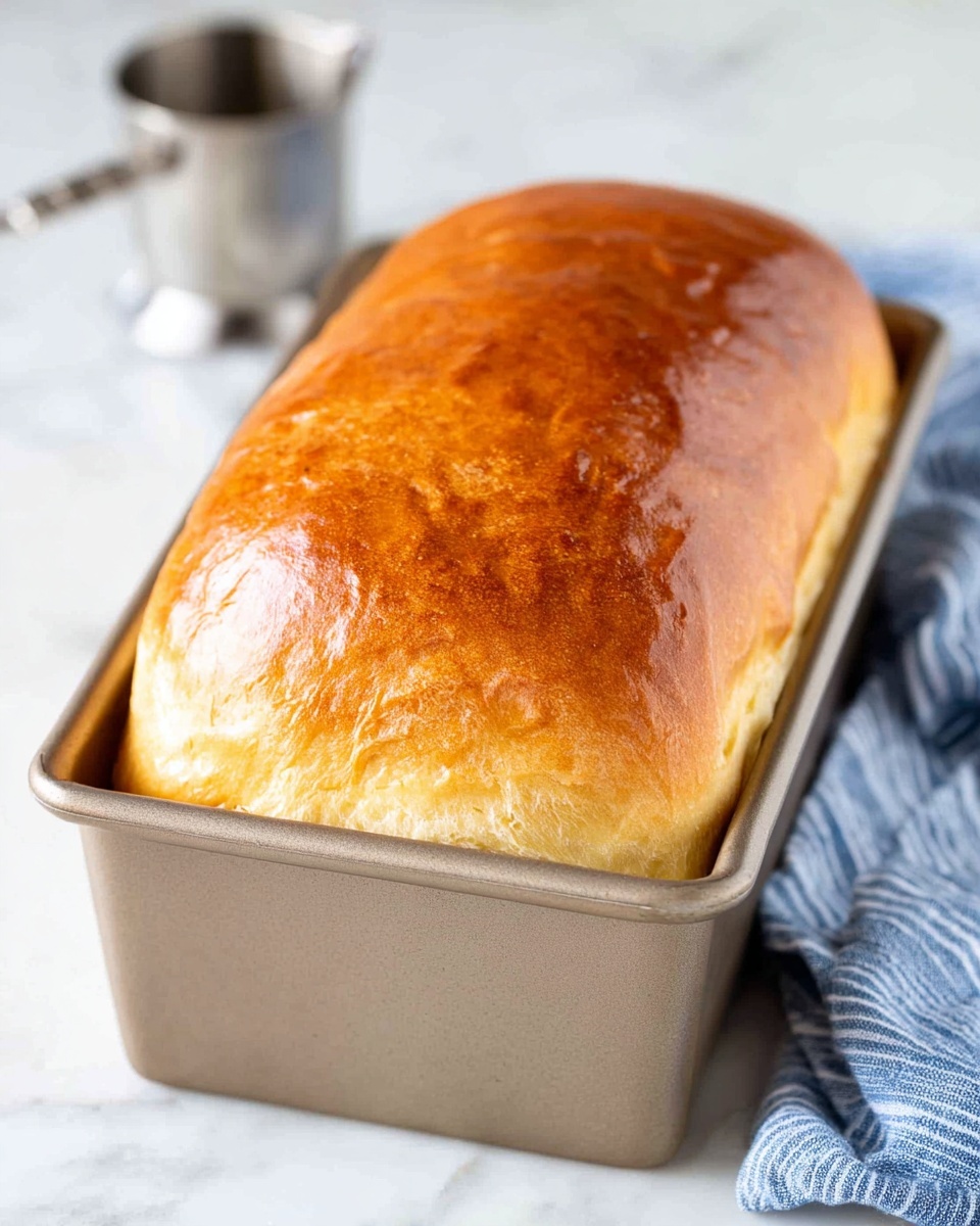 A loaf of golden brown bread with a shiny, smooth top crust rests inside a rectangular metal loaf pan. The bread looks soft and fluffy underneath the crust. In the background, there is a small metal measuring cup and a white marbled surface underneath everything. A blue and white striped cloth is seen near the pan, adding a soft touch to the image. photo taken with an iphone --ar 4:5 --v 7