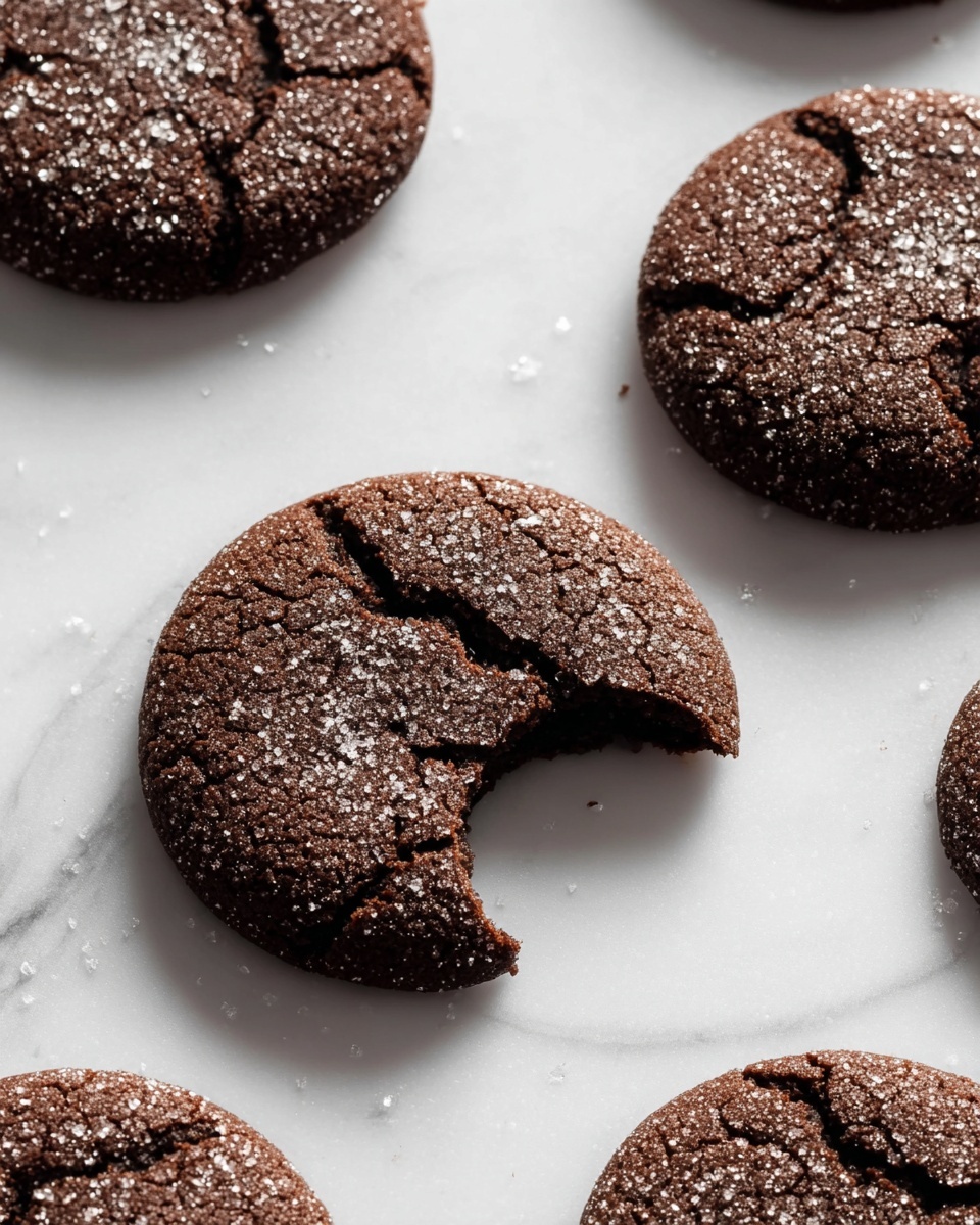 Several round chocolate cookies are placed on a white marbled surface, each cookie showing a rough cracked texture with sugar crystals sprinkled on top. One cookie near the center has a bite taken out of it, revealing the soft inside. The cookies are dark brown with a slightly matte finish and fine cracks. The scene is lit with soft natural light, highlighting the sugar sparkle and the cookie cracks. Photo taken with an iphone --ar 4:5 --v 7