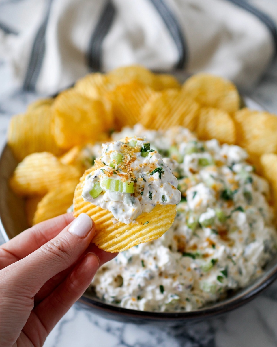 The dish shows a close-up of a creamy dip placed on a white plate sitting on a white marbled surface. The dip itself is textured, white with small green herb pieces mixed throughout, along with tiny light green and translucent vegetable pieces. It is topped with some fine brown crumbs and scattered bright green herb sprigs, giving it a fresh look. A light yellow ridged chip is dipped partially into the creamy mixture, adding contrast to the image. The setting includes a white and gray striped cloth napkin partially visible on the bottom right. photo taken with an iphone --ar 4:5 --v 7