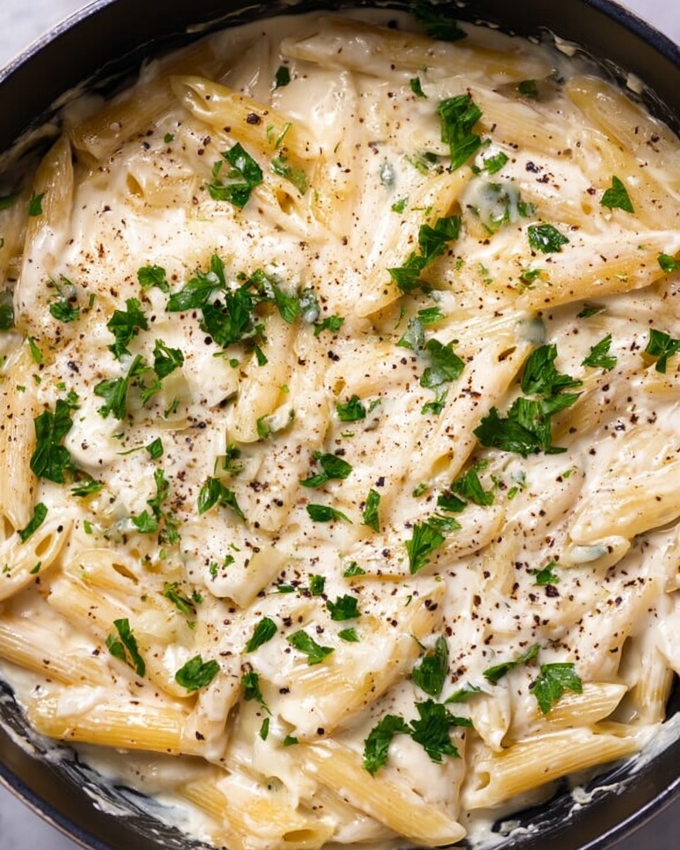 A close-up view of a pan filled with cooked penne pasta covered in a creamy white sauce. The sauce is thick and smooth, mixed well with the pasta, and sprinkled with fresh green parsley leaves scattered evenly on top. There are light white shavings of cheese spread over the pasta, with black pepper dots giving a speckled texture across the surface. The overall color mix is light beige from the creamy sauce, pale yellow from the pasta, bright green from the parsley, and touches of black from the pepper. The pan edges are dark, contrasting the colors inside. The dish is set on a white marbled surface photo taken with an iphone --ar 4:5 --v 7