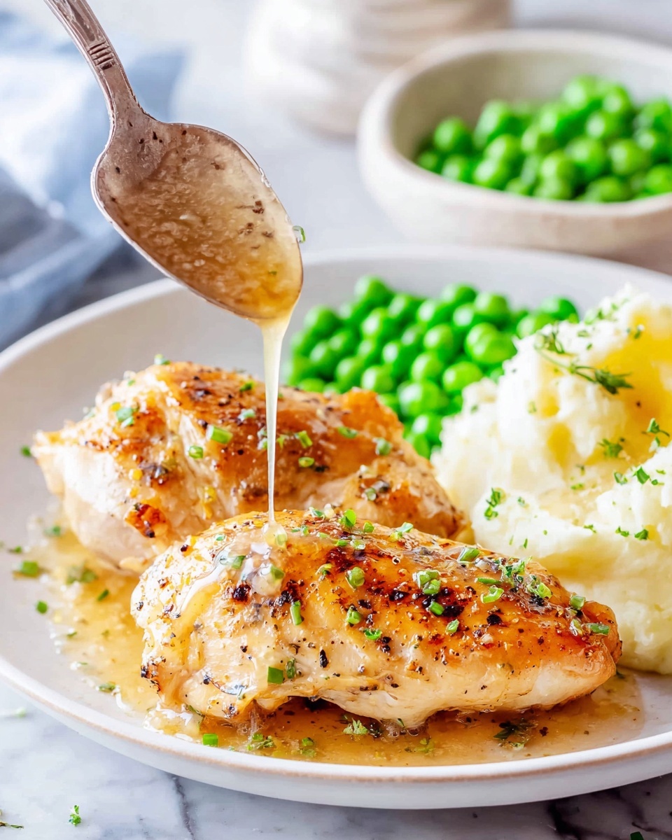 A white plate holds two grilled chicken fillets with a light golden brown color, speckled with small green herb pieces on top. Underneath, there is a smooth white layer of mashed potatoes, slightly visible around the edges. To the back right side of the plate, there is a small pile of bright green peas. A woman's hand holds a silver spoon above the chicken, pouring a light brown sauce with small bits over the fillets. The background features a white marbled surface and blurred lemons and a bowl of peas can be seen. Photo taken with an iphone --ar 4:5 --v 7