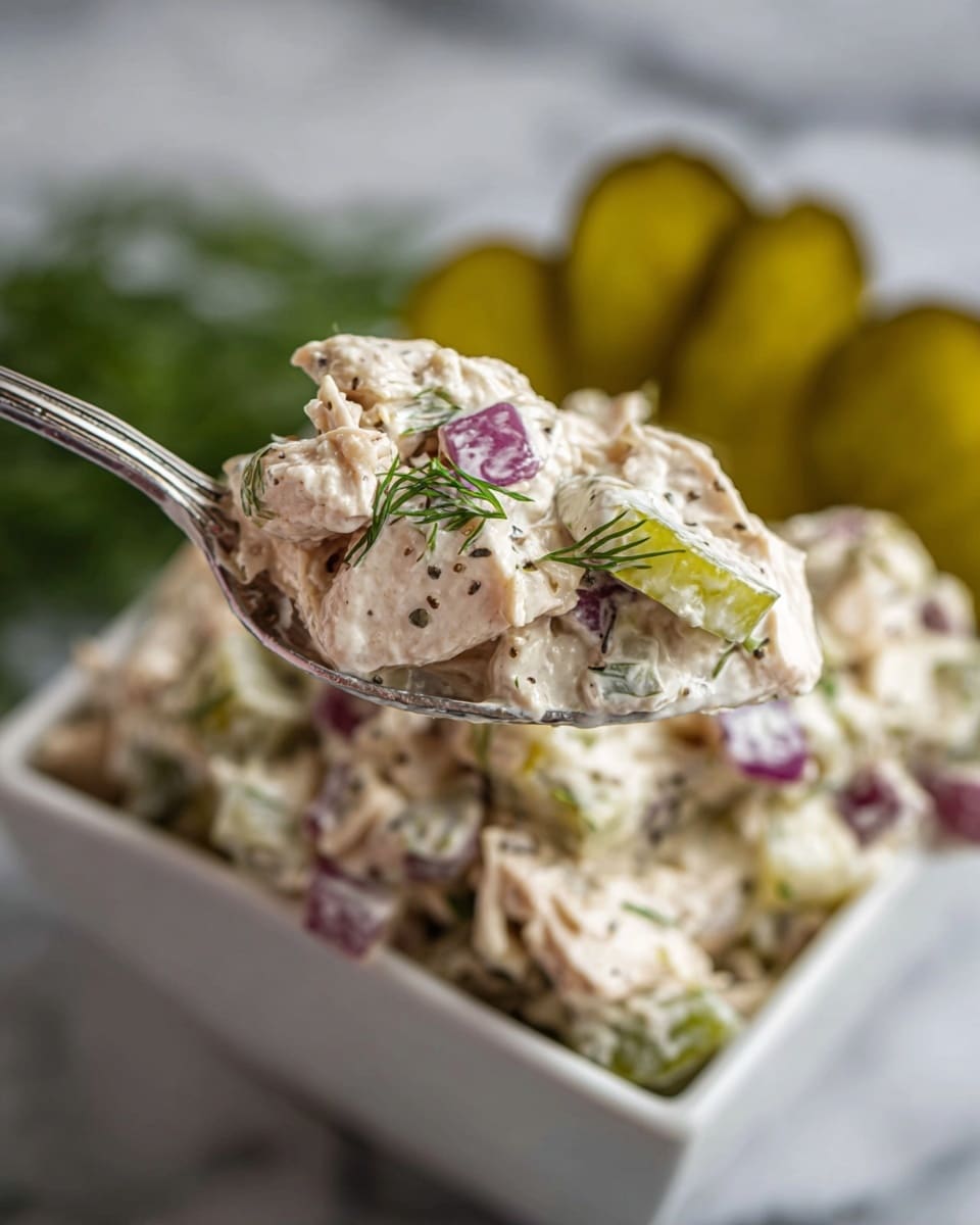 A white bowl filled with a creamy mixture made of shredded light beige chicken layered with small green celery pieces and diced pickles, all coated evenly in a white creamy sauce with flecks of black pepper and small green dill leaves scattered throughout; a wooden spoon is resting on the right rim of the bowl, slightly submerged in the mixture, and a small sprig of fresh green dill floats on top in the center; around the bowl on a white marbled surface sit sliced pickles and a green cloth. photo taken with an iphone --ar 4:5 --v 7