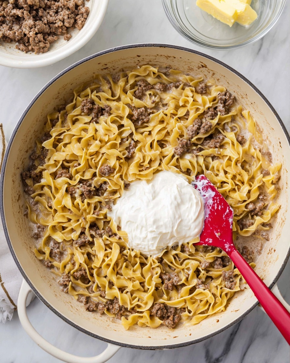 A large white pan filled with cooked yellow egg noodles and browned ground meat mixed together in a light creamy sauce. On top in the center, there is a dollop of thick white cream, partially mixed around a red spatula resting inside the pan on the right side. The pan sits on a white marbled surface with part of two bowls visible at the top edge—one white bowl with bits of ground meat and one clear glass bowl with a small piece of yellow butter. photo taken with an iphone --ar 4:5 --v 7