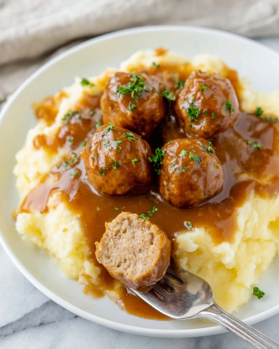 Slow Cooker Salisbury Steak Meatballs Recipe 4 A white plate shows a layer of creamy mashed potatoes at the bottom with a soft texture and light yellow color. On top, there are five round meatballs covered in thick, shiny brown gravy that glistens under the light. The meatballs have a smooth and slightly bumpy surface. Small green parsley pieces are sprinkled on the gravy for color contrast. In the front, a meatball is sliced in half on a silver fork, showing a dense, light brown inside. The plate rests on a white marbled surface. photo taken with an iphone --ar 4:5 --v 7