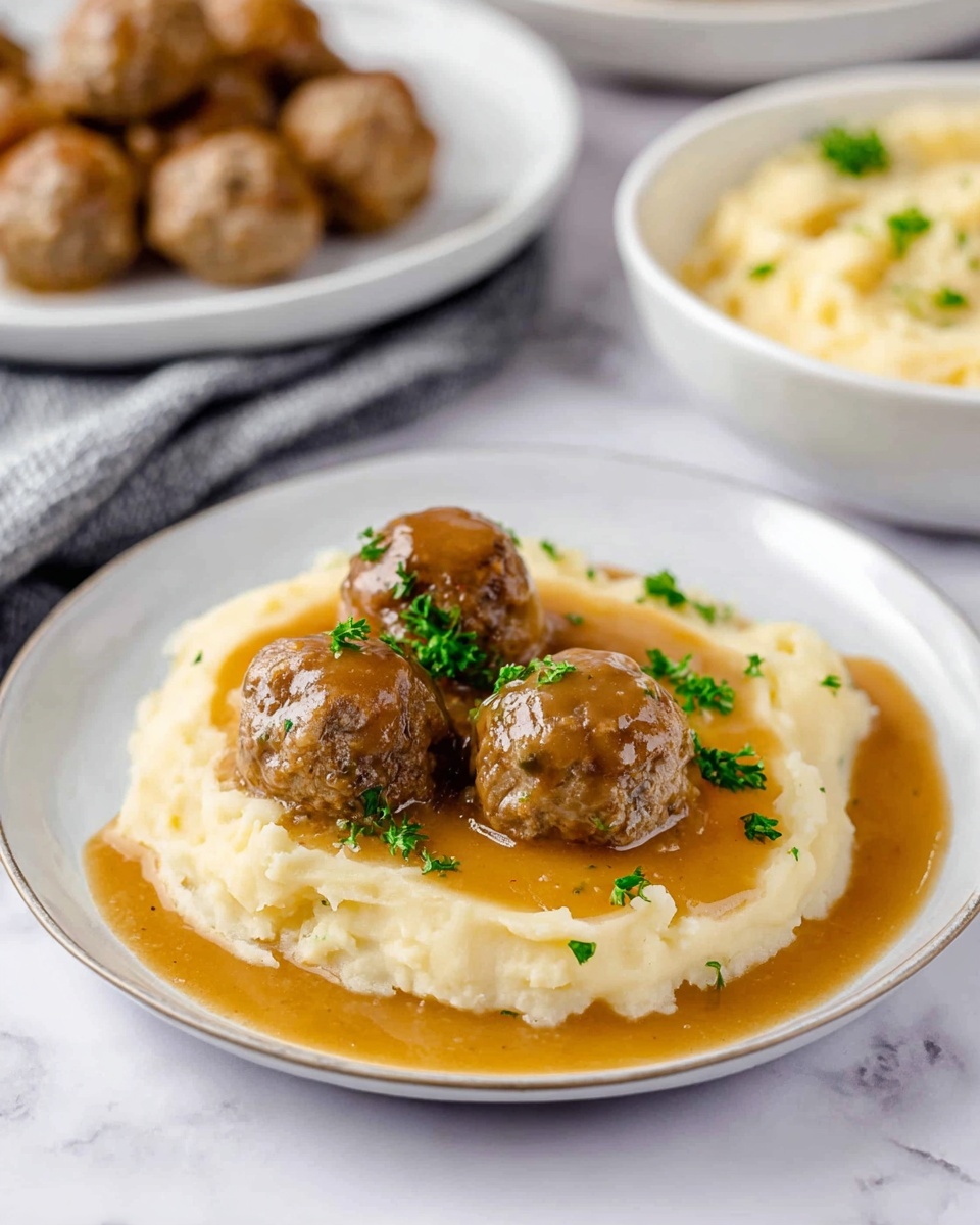 The image shows a white plate with a thick, creamy layer of mashed potatoes spread evenly across most of the plate. On top of the mashed potatoes, there are three brown meatballs covered with a shiny brown gravy that pools slightly around them. Small green parsley pieces are sprinkled over the meatballs and gravy, adding some color contrast. In the background, another white plate holds additional meatballs, and there is a bowl containing more mashed potatoes. The setup is on a white marbled surface. The photo taken with an iphone --ar 4:5 --v 7