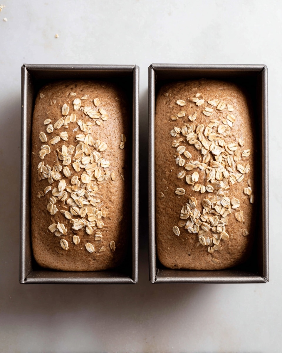 Two dark brown bread dough loaves are shown in metal loaf pans side by side, each loaf sprinkled with light beige rolled oats on top. The dough has a smooth but slightly textured surface, filling most of the pan's space and rising slightly above the edges. The pans rest on a white marbled surface with soft natural light highlighting the texture of the dough and oats. photo taken with an iphone --ar 4:5 --v 7
