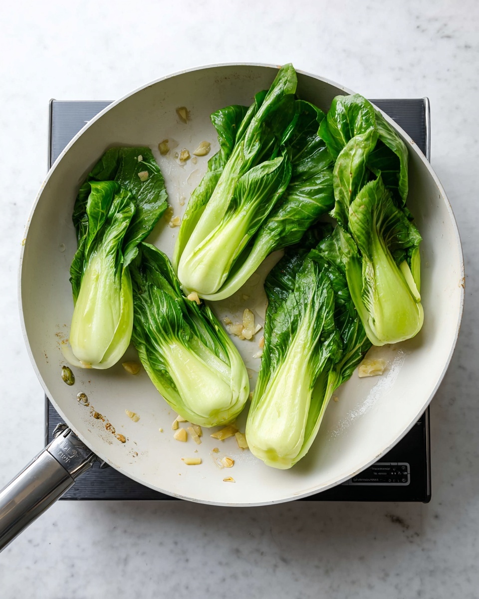 A white pan sits on a white marbled surface, containing four pieces of bok choy arranged in a rough circle. Each bok choy has pale green thick stalks at the bottom and bright green leafy tops that are slightly wilted. The pan also has small bits of garlic scattered around the bok choy. A silver handle extends from the pan toward the bottom left corner, with a black cooking device partly visible underneath the pan on the right side. The light shines softly from above, highlighting the fresh texture and colors of the bok choy. photo taken with an iphone --ar 4:5 --v 7