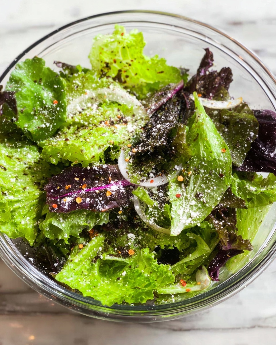 A clear glass bowl filled with mixed leafy greens, including bright green and deep purple lettuce leaves, layered roughly with varied sizes and fresh textures. Finely grated white cheese lightly dusts the top, sprinkled evenly over the greens along with small red pepper flakes adding spots of red color. Thin rings of pale onion are scattered sparsely on top. The bowl sits on a white marbled surface. photo taken with an iphone --ar 4:5 --v 7