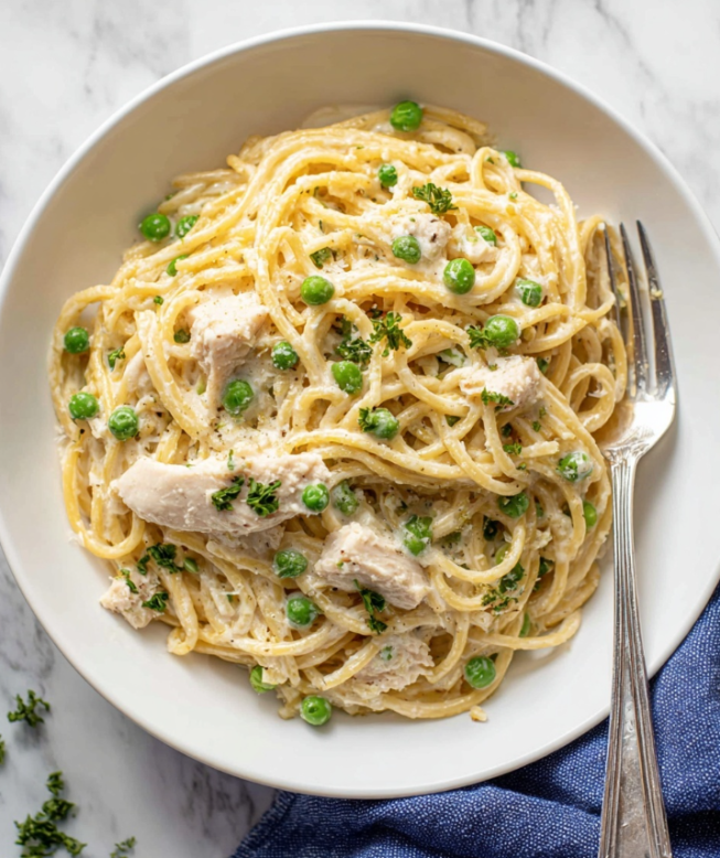 A white bowl holds a creamy pasta dish with three main layers. The bottom layer shows thick, light yellow spaghetti noodles, arranged loosely in mostly circular shapes. Mixed within the noodles are small bits of light beige chicken pieces and bright green peas scattered evenly. The top layer is a smooth, off-white creamy sauce coating the pasta and chicken, with tiny green parsley pieces sprinkled on top for color. A vintage silver fork rests inside the bowl on the right side, while the bowl sits on a white marbled surface with a folded blue cloth nearby. Photo taken with an iphone --ar 4:5 --v 7