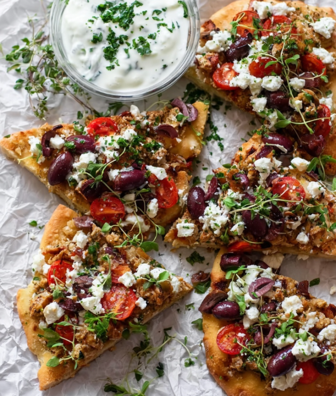 The image shows several triangular slices of Greek flatbread arranged on crumpled white paper over a white marbled surface. Each flatbread slice has a golden-brown base and is topped with layers starting with crumbled light beige meat, bright red tomato slices, and white crumbled feta cheese. On top, there are dark purple olive pieces spread across, along with scattered small green herb leaves and sprouts, adding color and freshness. To the side, there is a clear bowl with creamy white tzatziki sauce garnished with finely chopped green herbs. The setting is bright and fresh with a rustic and wholesome vibe. Photo taken with an iphone --ar 4:5 --v 7
