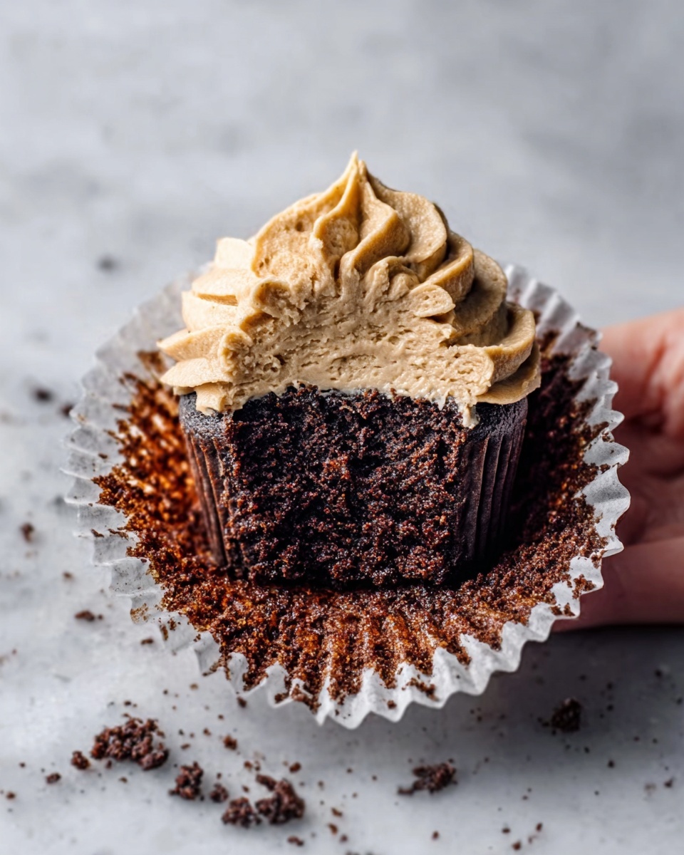 A close-up of a chocolate cupcake with a thick layer of light brown frosting swirled on top, slightly eaten from the side showing the moist dark brown cake inside. The cupcake is in a white paper liner, resting on a white marbled surface with scattered chocolate crumbs around. A woman's hand is gently holding the cupcake from the side. Photo taken with an iphone --ar 4:5 --v 7