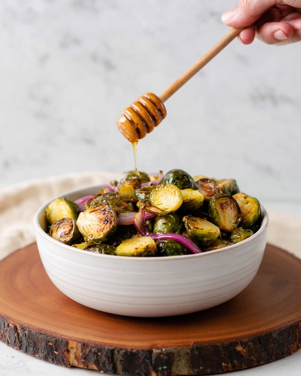A white bowl full of halved roasted Brussels sprouts, showing golden brown edges and some pieces of cooked red onion mixed in, is placed on a round wooden board. A woman's hand holds a wooden honey dipper above the bowl, with honey slowly dripping down onto the vegetables. The background and surface have a white marbled texture, giving a clean and bright look. photo taken with an iphone --ar 4:5 --v 7