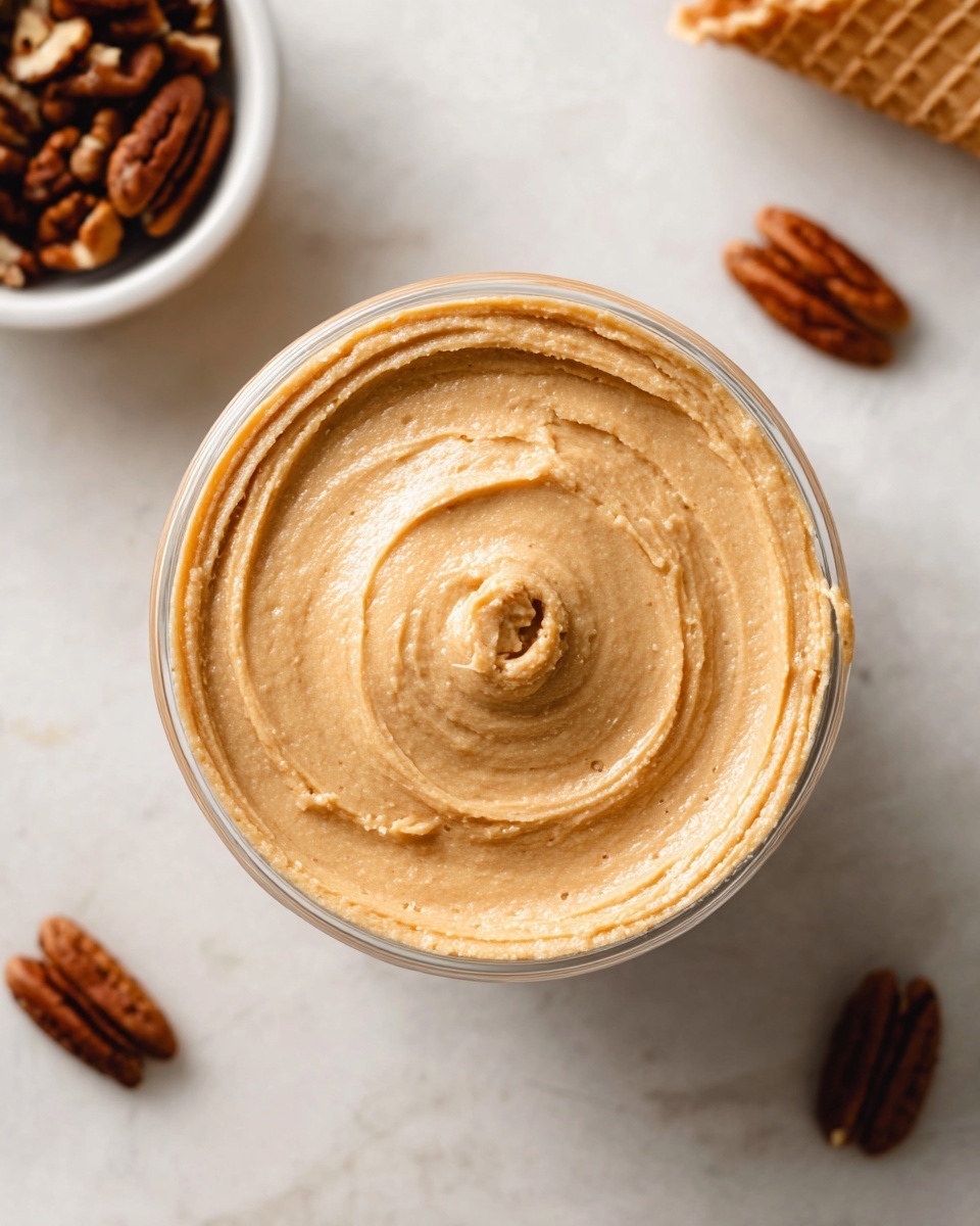 A clear glass bowl filled with one creamy layer of light brown peanut butter, smoothly swirled in a circular pattern with a small indentation in the center. The bowl sits on a white marbled surface. Nearby, there is a white bowl with pecans and two waffle cones placed diagonally, one on the top left and one on the bottom right edges of the image. The lighting is soft and natural, highlighting the smooth texture of the peanut butter. photo taken with an iphone --ar 4:5 --v 7
