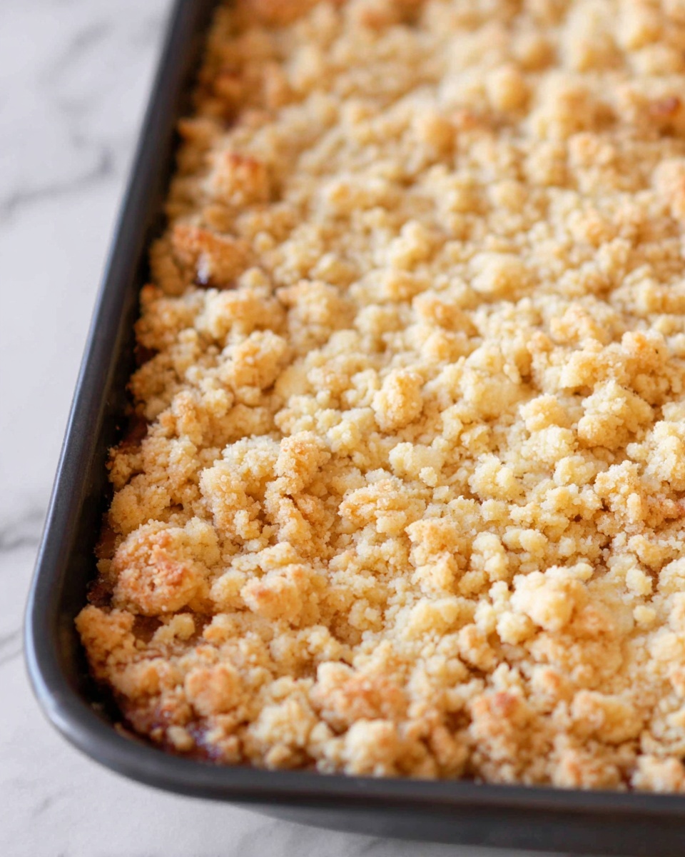 A close-up image of a baked crumb topping with one layer, showing small, uneven crumbles in a light golden and cream color, with a slightly crispy texture. The crumbs are spread thickly and cover the entire visible surface in a baking pan with dark edges, all placed on a white marbled texture. The photo taken with an iphone --ar 4:5 --v 7