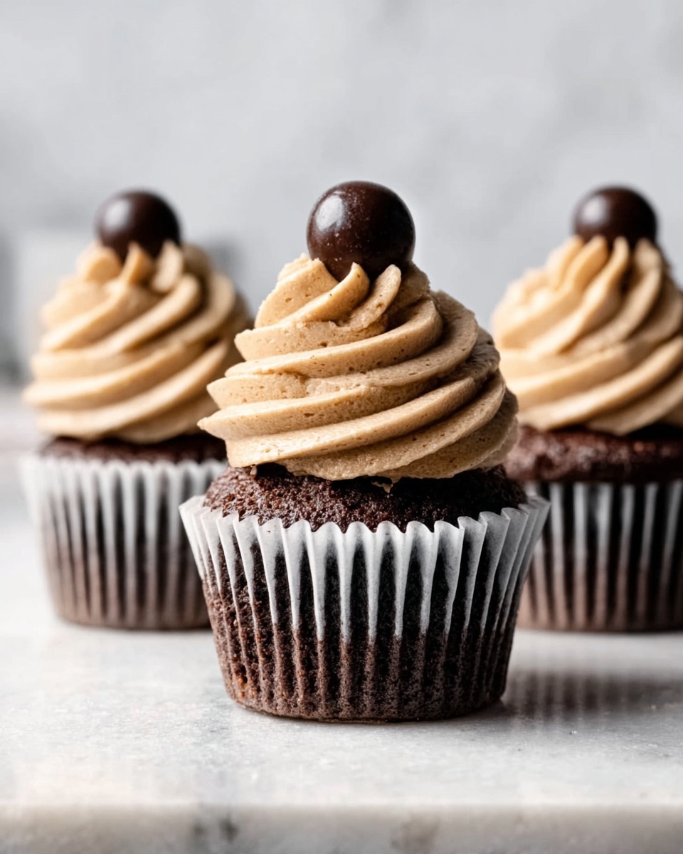 The image shows three chocolate cupcakes lined up on a white marbled surface, each in white and dark brown striped paper wrappers. Each cupcake has one thick, tall swirl of light brown frosting on top, with a smooth, creamy texture and ridges from piping. A small, round, dark chocolate ball sits on the top center of the frosting swirl on each cupcake. The background is blurred with soft white tones, making the cupcakes the main focus. The photo taken with an iphone --ar 4:5 --v 7