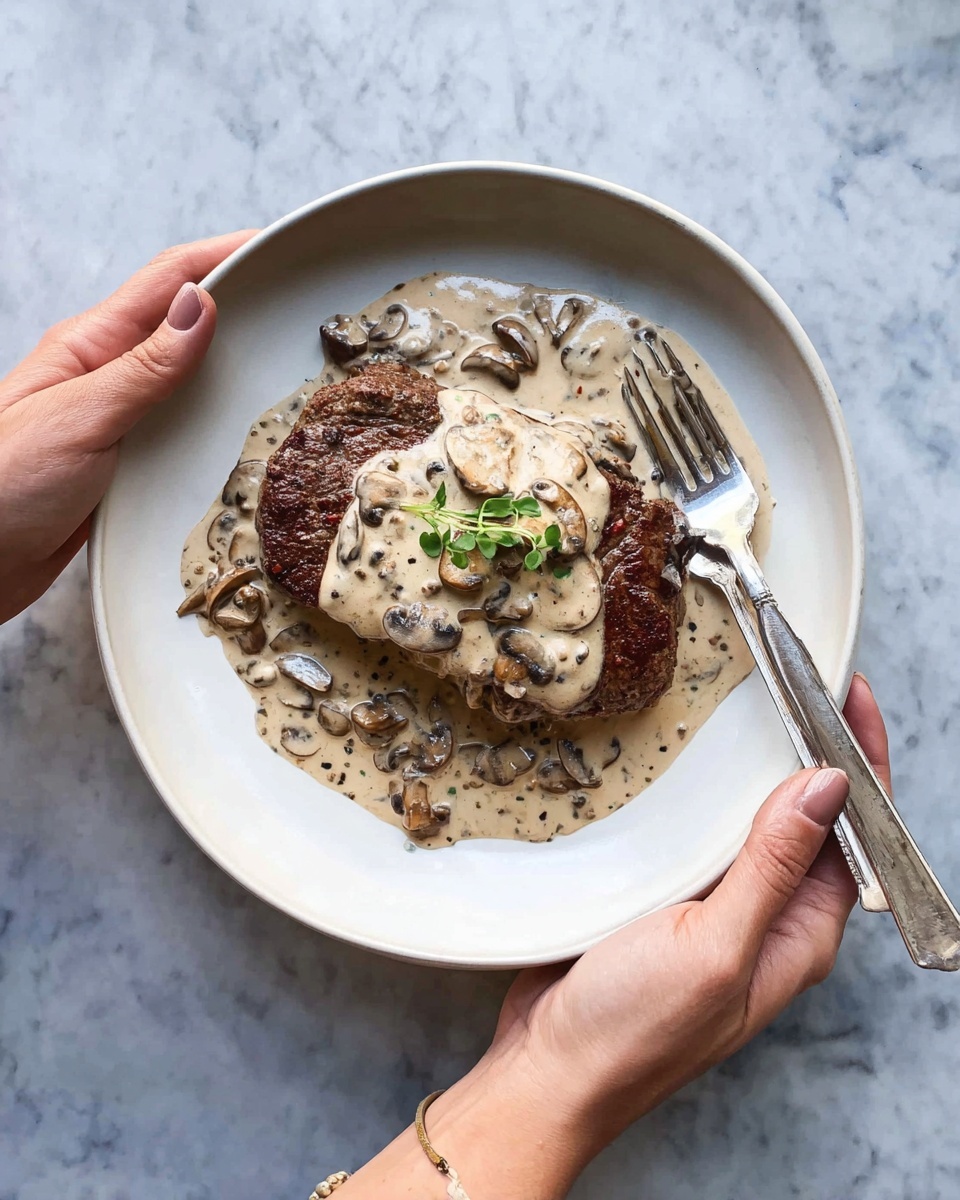 A white plate is held by two woman's hands, containing one thick, browned steak as the base layer with a rich creamy mushroom sauce on top. The sauce has visible sliced mushrooms and small black pepper specks, spreading from the center to the edges of the steak and plate. A small green herb garnish is placed in the center on the sauce. A silver fork rests on the plate partly submerged in the sauce near the steak. The background shows a white marbled texture. photo taken with an iphone --ar 4:5 --v 7
