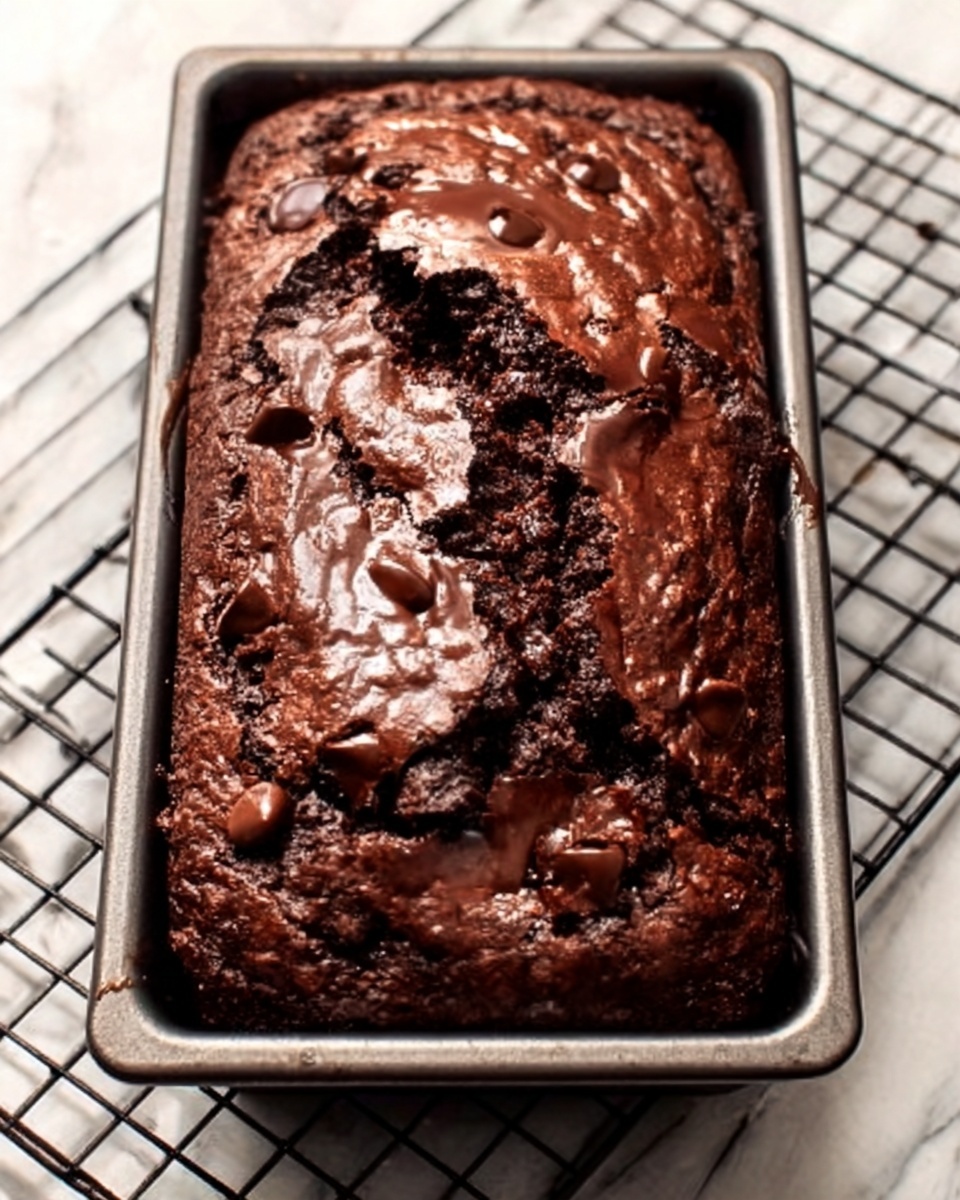 A loaf of rich chocolate brownie sits in a rectangular metal pan on a white marbled surface. The brownie has a cracked, shiny top showing melted chocolate chips beneath the crust. The texture looks dense and moist with visible chunks of chocolate inside. The pan is surrounded by a black wire cooling rack. photo taken with an iphone --ar 4:5 --v 7