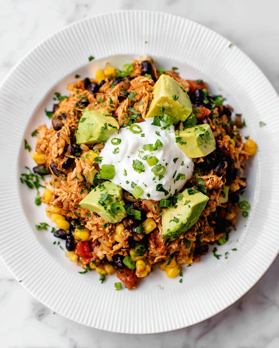 A white ridged plate holds a colorful layered dish starting with a base of shredded brownish-orange chicken mixed with black beans and yellow corn. On top of this are scattered bright green avocado chunks and chopped green onion slices. The dish is garnished with finely chopped fresh herbs and has a dollop of smooth white sour cream sitting in the center. The plate rests on a white marbled surface. photo taken with an iphone --ar 4:5 --v 7