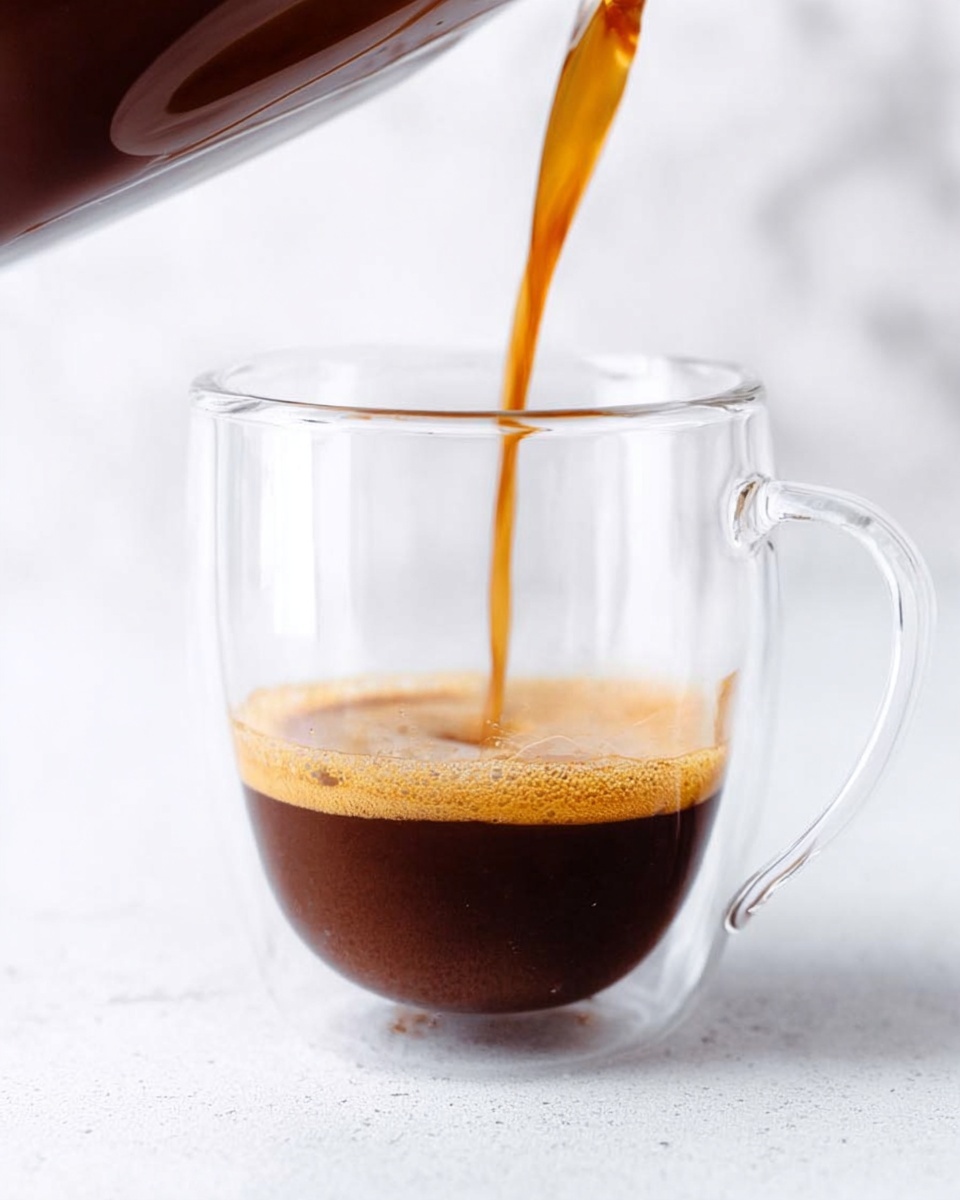 A clear glass cup with a handle is shown, positioned in front of a white marbled texture background. Dark brown liquid coffee is being poured from a glass container into the cup, creating a thin stream that touches the bottom where a small layer of coffee is already resting, with a slight light brown foam forming on top. The scene is bright and simple, focusing on the clarity of the glass and the rich color of the coffee as it fills the empty space in the cup. photo taken with an iphone --ar 4:5 --v 7