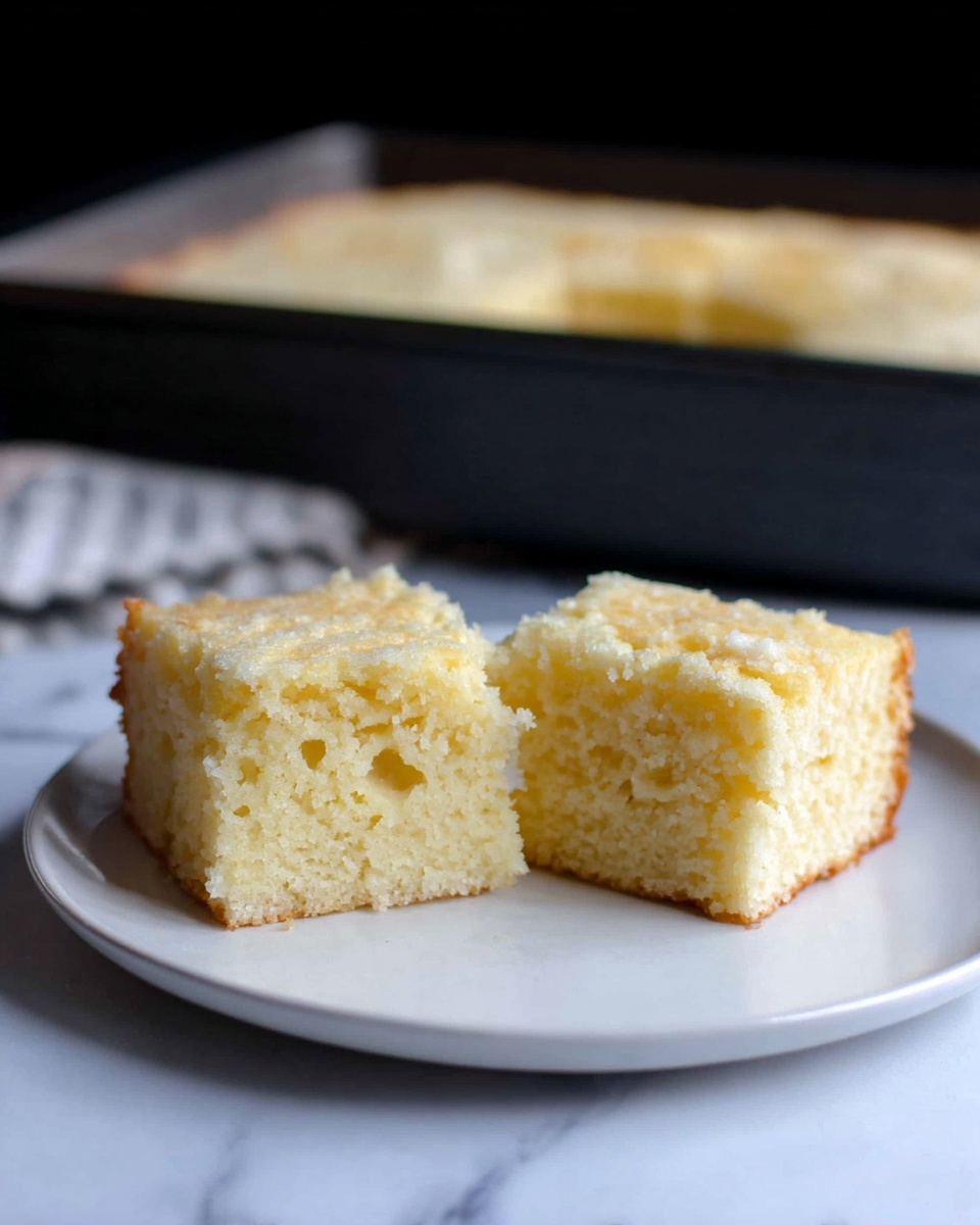 Two square pieces of light yellow cake sit side by side on a white plate, each piece showing a soft and moist texture with small air holes inside. The cake has a crumbly top layer that looks slightly lighter in color. The plate is placed on a white marbled surface, and in the background, there is a dark baking pan filled with more of the same cake. The lighting highlights the soft texture and crumbly top of the cake. photo taken with an iphone --ar 4:5 --v 7