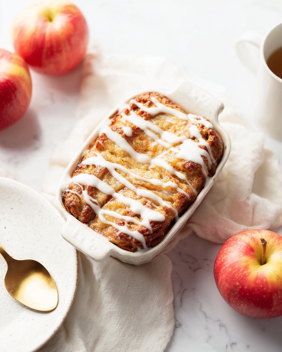 A small white baking dish holds a golden-brown baked dessert with a slightly rough and crumbly texture on top. The top is drizzled unevenly with white icing that contrasts with the warm color below. The dish sits on a soft white cloth laid over a white marbled surface. Nearby are two red apples with a shiny, smooth skin showing red and yellow hues, and a white cup and a white plate with a golden spoon resting beside it. The lighting highlights the warm colors and soft textures in the scene, creating a cozy and inviting feeling. photo taken with an iphone --ar 4:5 --v 7