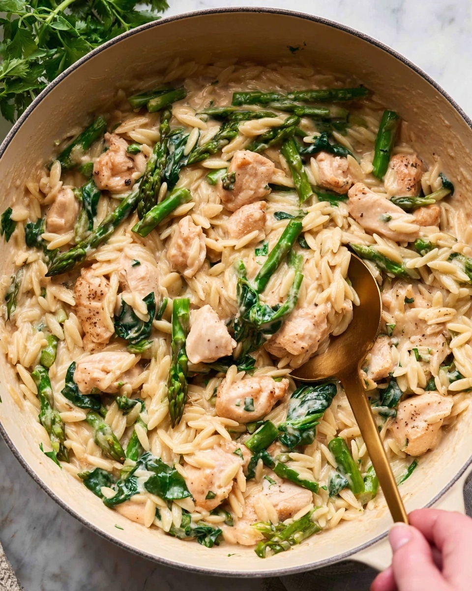 A close-up view of a cream-colored pan filled with a creamy pasta dish. The dish has three main layers: plump, tender pieces of light brown chicken scattered evenly; thick, short grain orzo pasta coated in a glossy, pale creamy sauce; and bright green asparagus tips and leafy spinach mixed throughout. A shiny gold spoon is inserted into the dish, held by a woman's hand at the back edge of the pan. The background shows a hint of green herbs on a white marbled surface. photo taken with an iphone --ar 4:5 --v 7